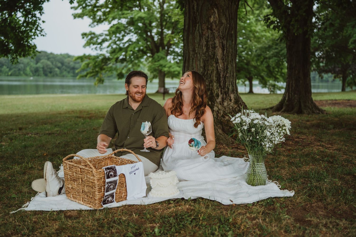 Couple enjoying a romantic picnic under a tree by a lake, sitting on a white blanket with a wicker basket and flowers.