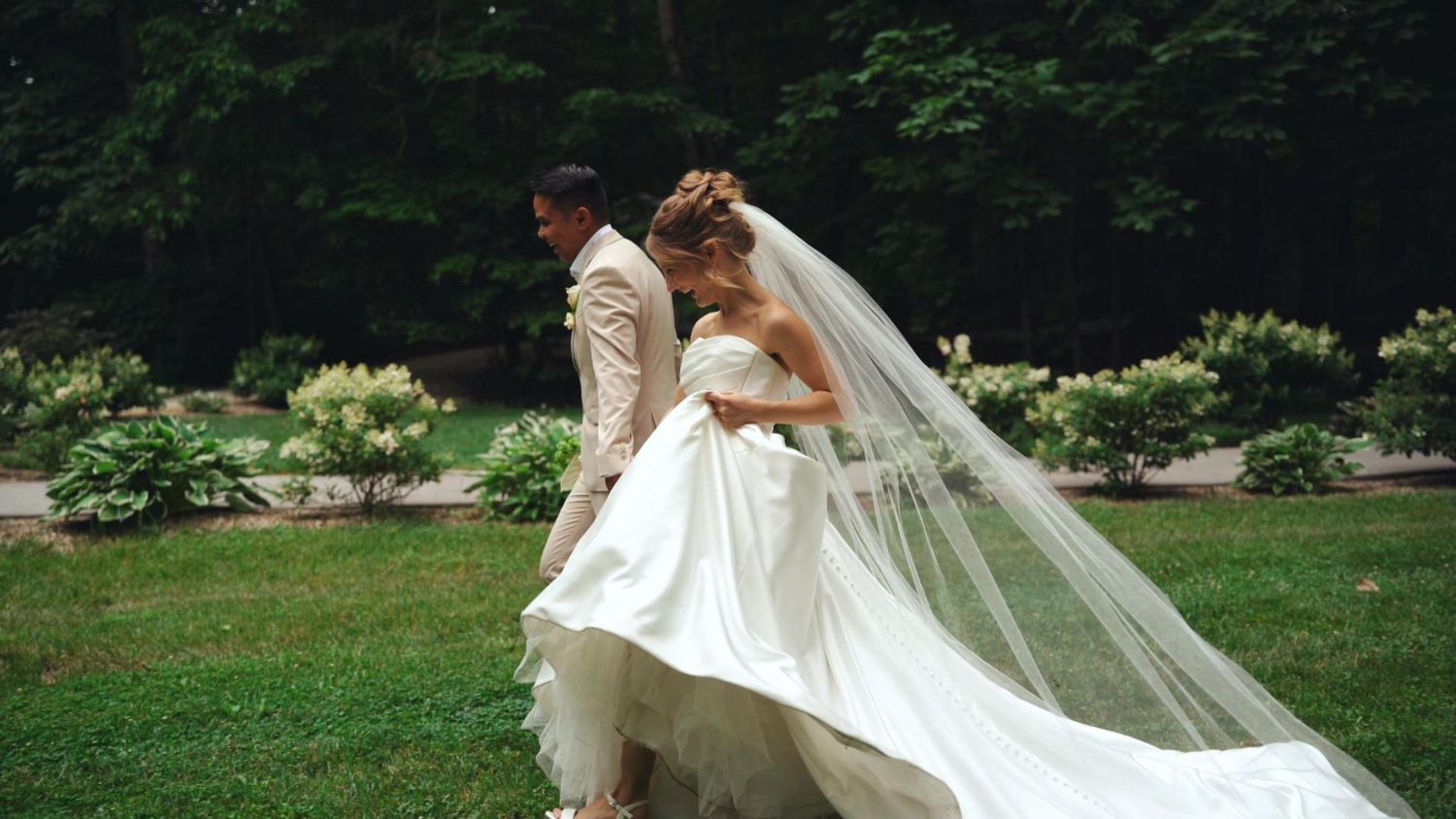 A bride in a white gown and veil walks with a groom in a tan suit on a lush green lawn.