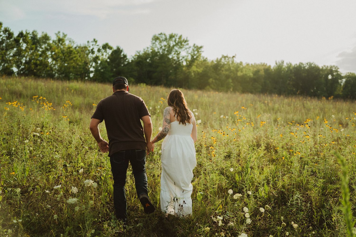 Couple holding hands walking through a sunlit wildflower meadow, woman in white dress, golden hour photography.