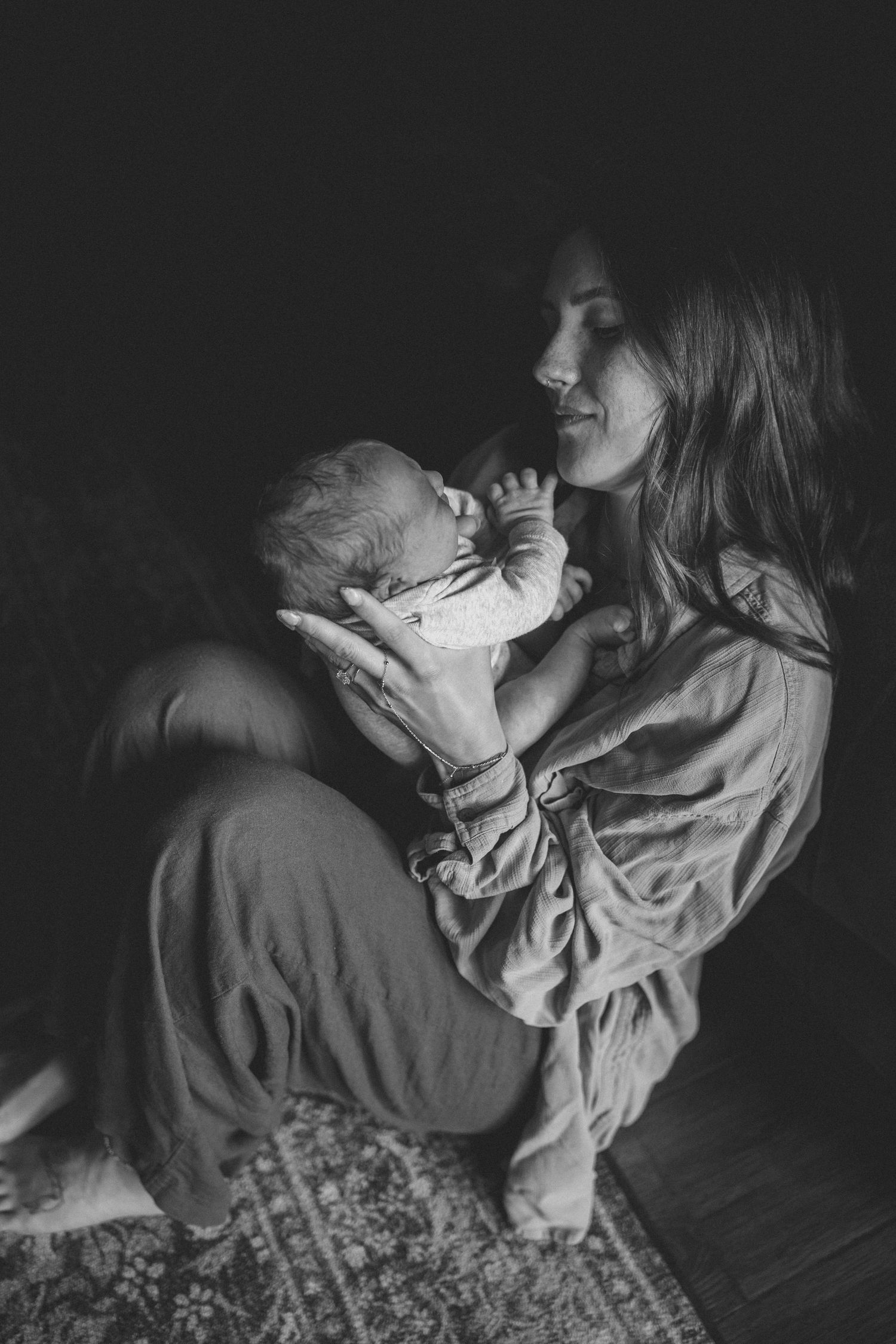 Black and white photo of a mother tenderly holding her newborn baby while sitting on a rug in soft natural light.