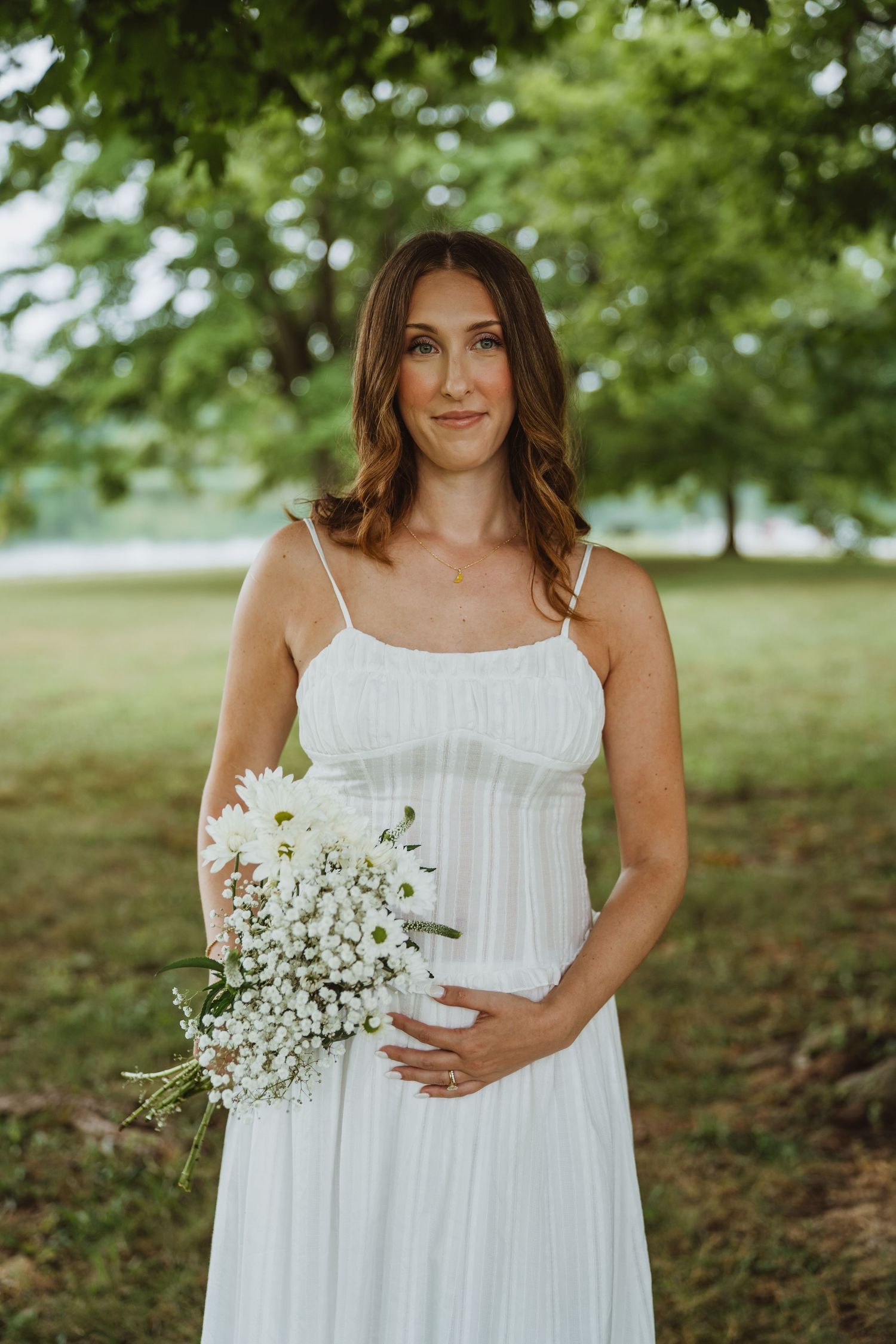 Pregnant woman in a white sundress holding a bouquet of white daisies under a large green tree.
