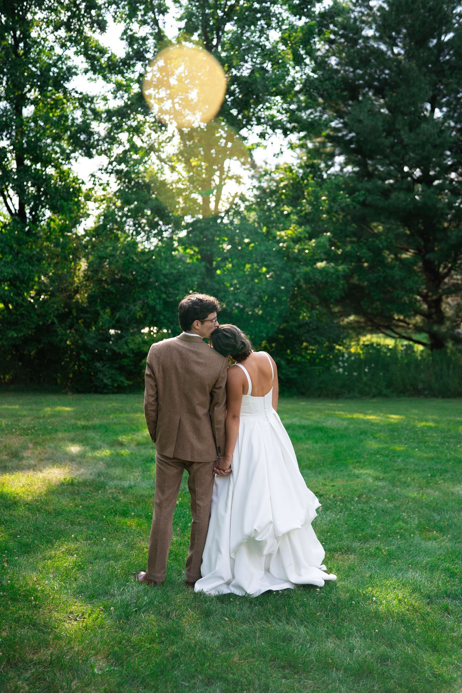Bride and groom standing hand in hand in lush green garden, viewed from behind, wearing white gown and brown suit.