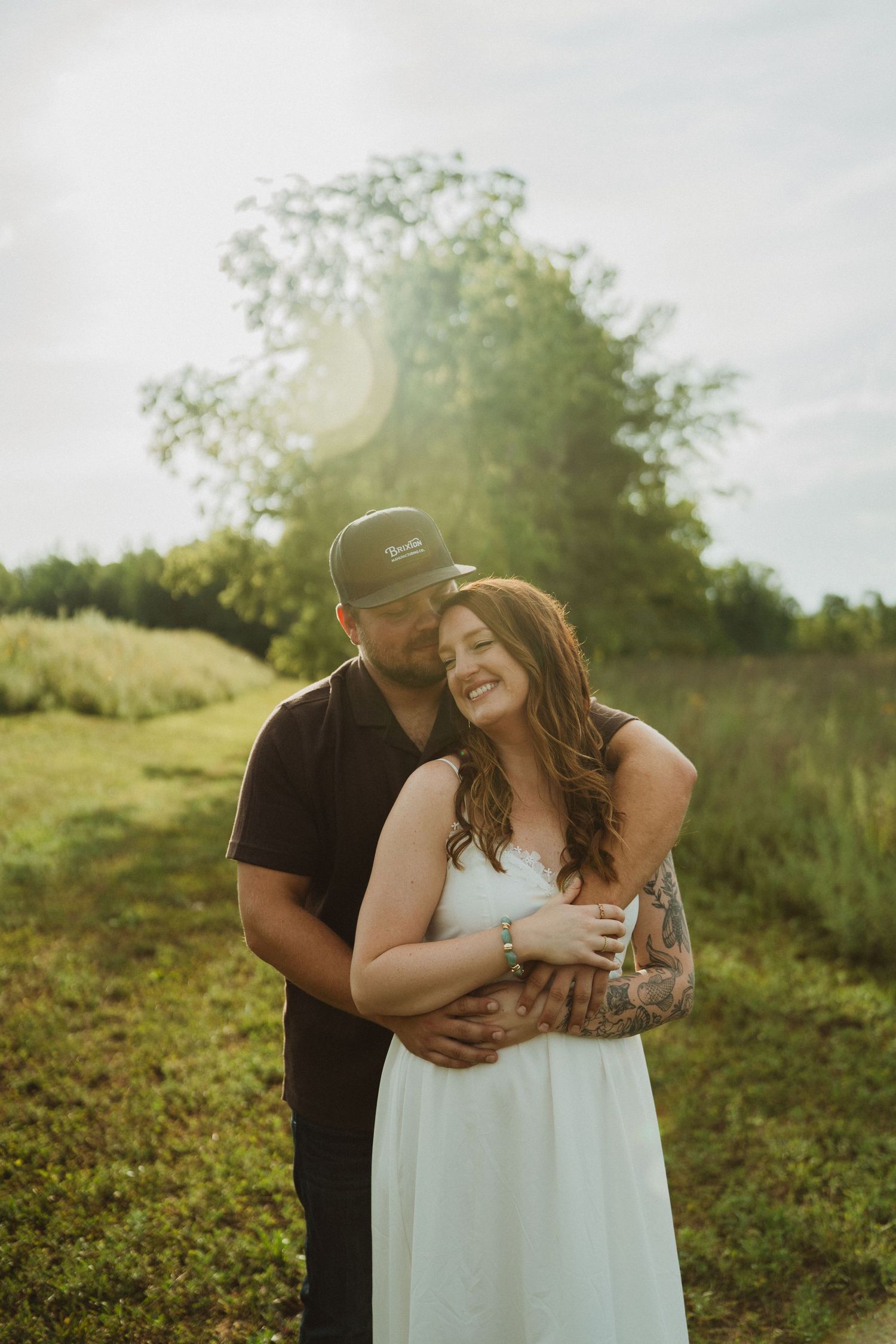 Happy couple cuddling in a sunlit meadow, woman with tattoos in white dress laughing while man holds her from behind.