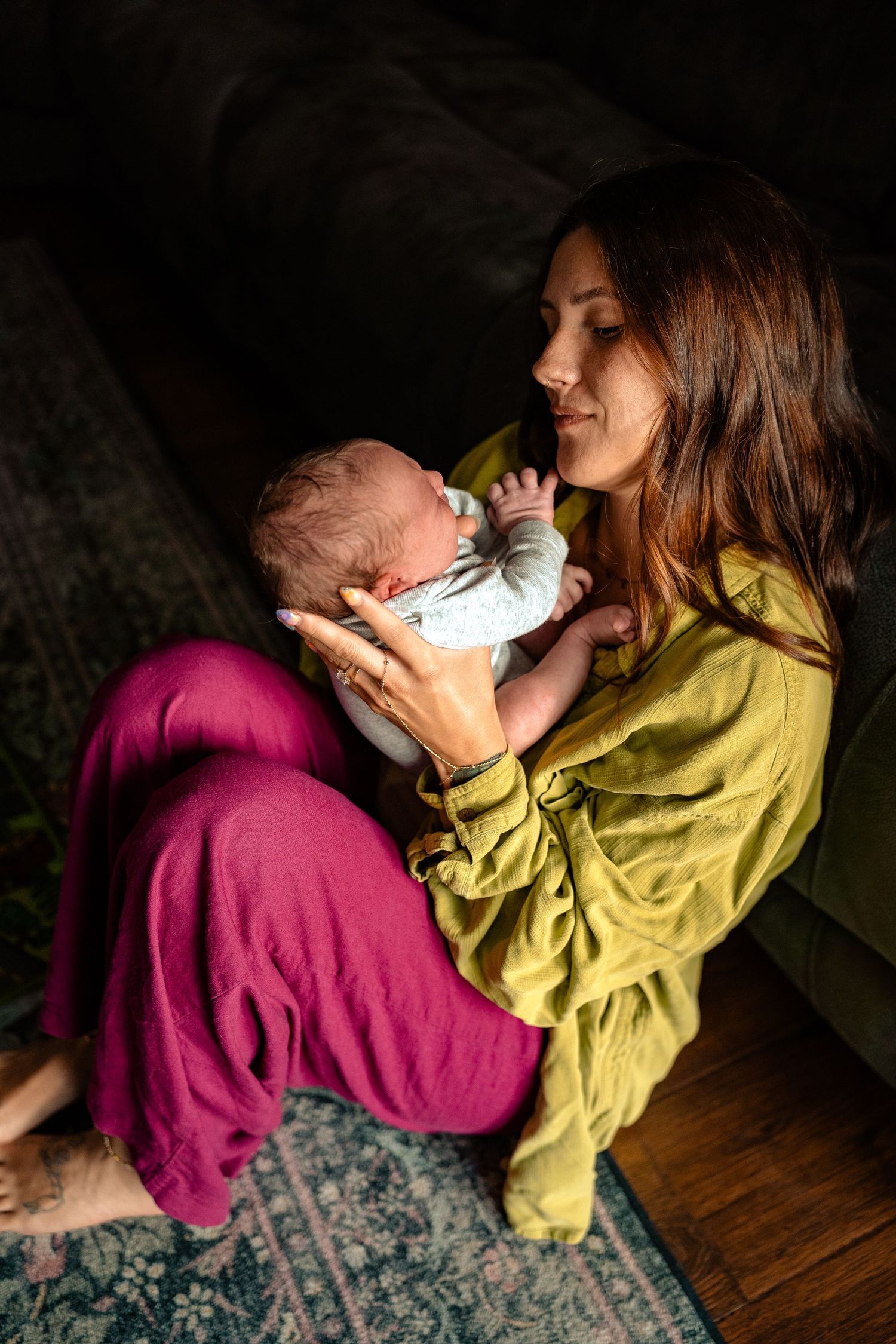 Mother in yellow-green blouse cradles sleeping newborn baby while seated on floor near dark sofa.