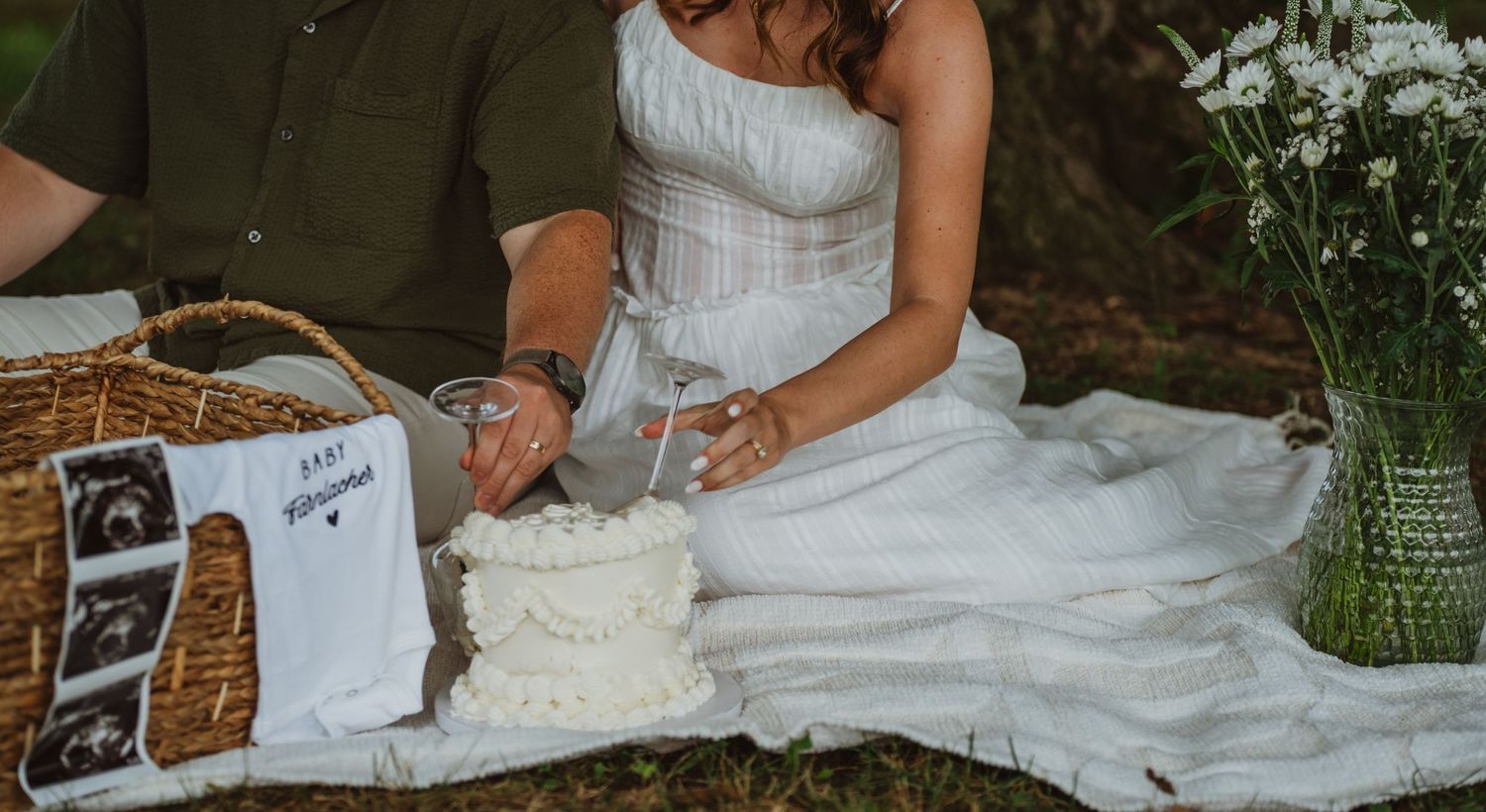Couple enjoying a romantic outdoor picnic with baby announcement items, ultrasound photos, and a white cake on a blanket.