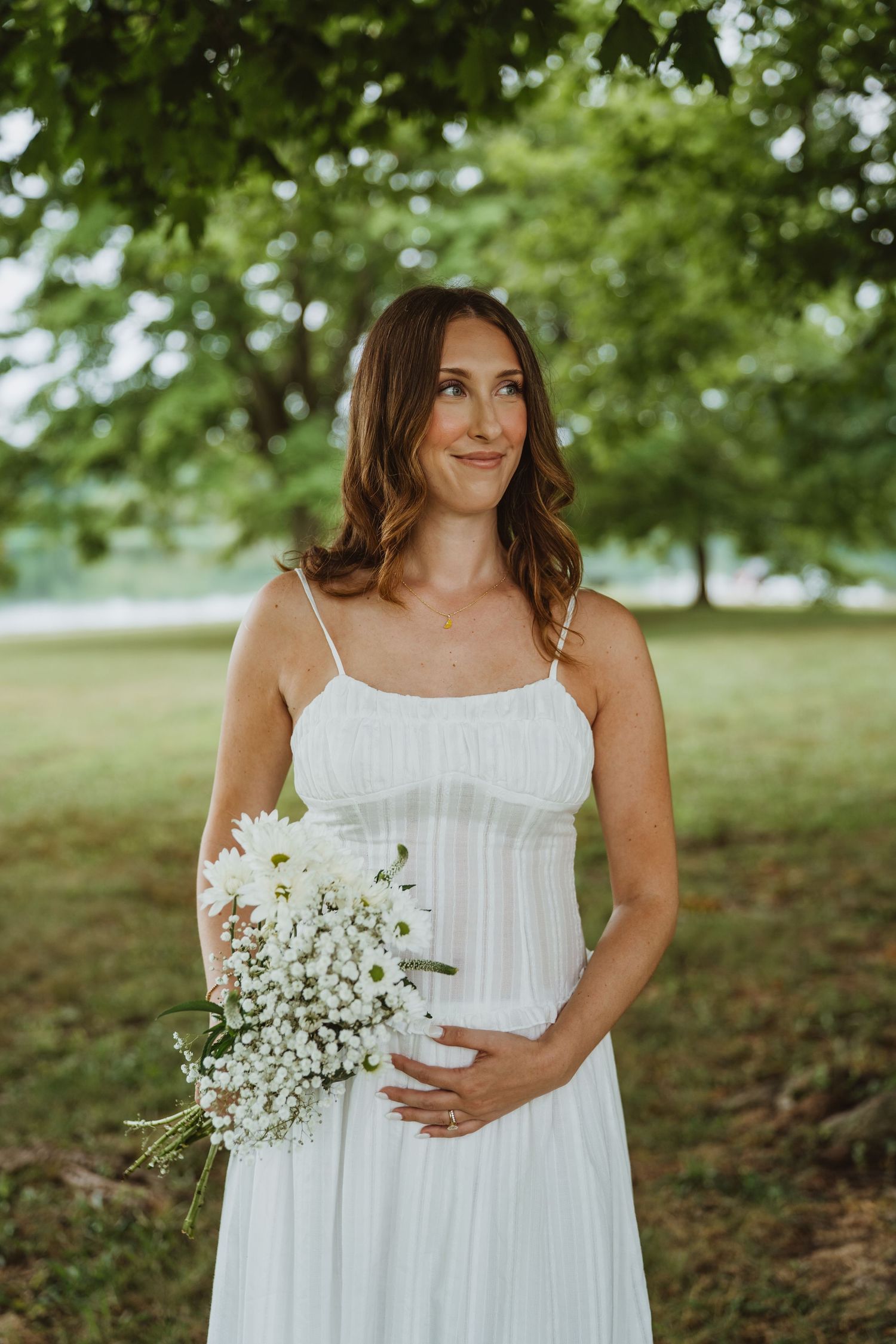 Smiling pregnant woman in white sundress holding white wildflower bouquet while cradling baby bump outdoors.