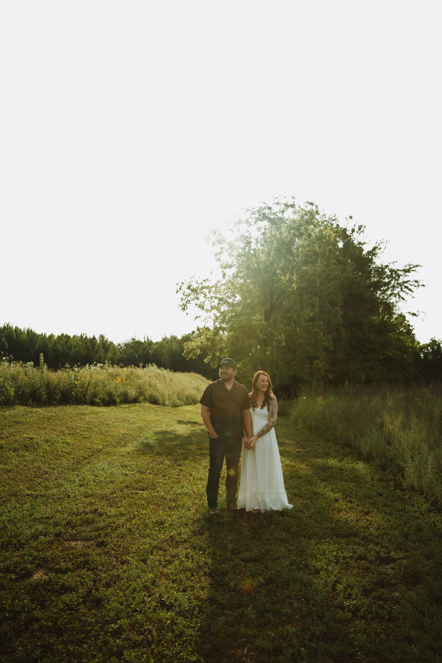 Couple in white dress and dark shirt holding hands in a sunlit green field with trees at golden hour.