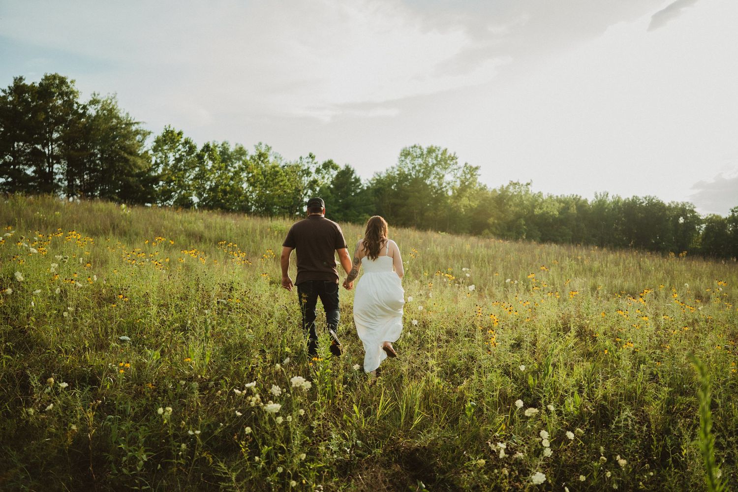 Couple strolling hand in hand through a lush wildflower field at sunset, woman wearing flowing white dress.