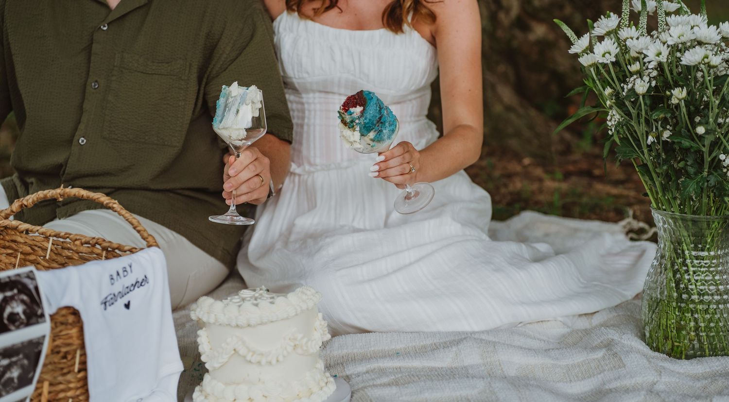 Couple in wedding attire holding decorated wine glasses beside a white cake and floral arrangement outdoors.