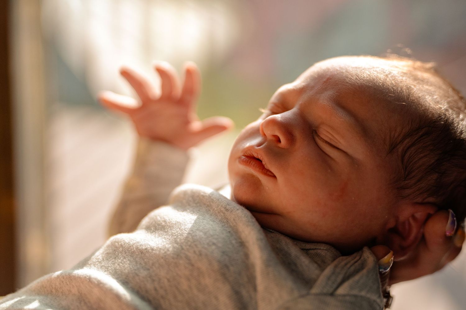 Peaceful newborn baby sleeping in warm sunlight, wearing a gray onesie with tiny hand raised.