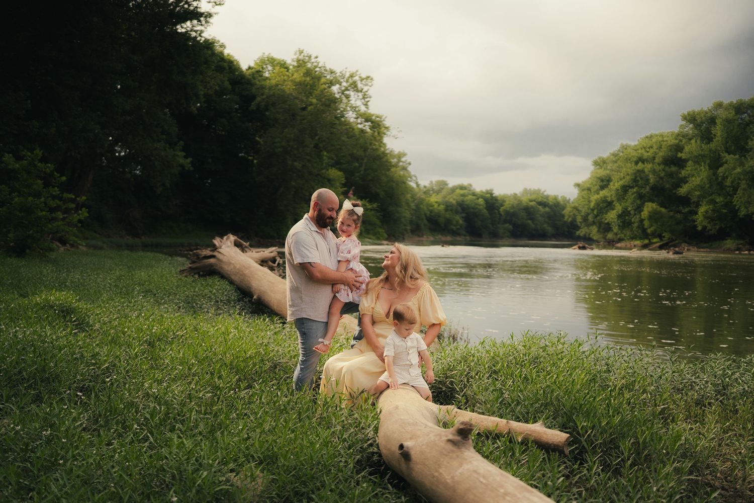 Family posing by a river on green grass with fallen logs and lush trees in a moody outdoor portrait.