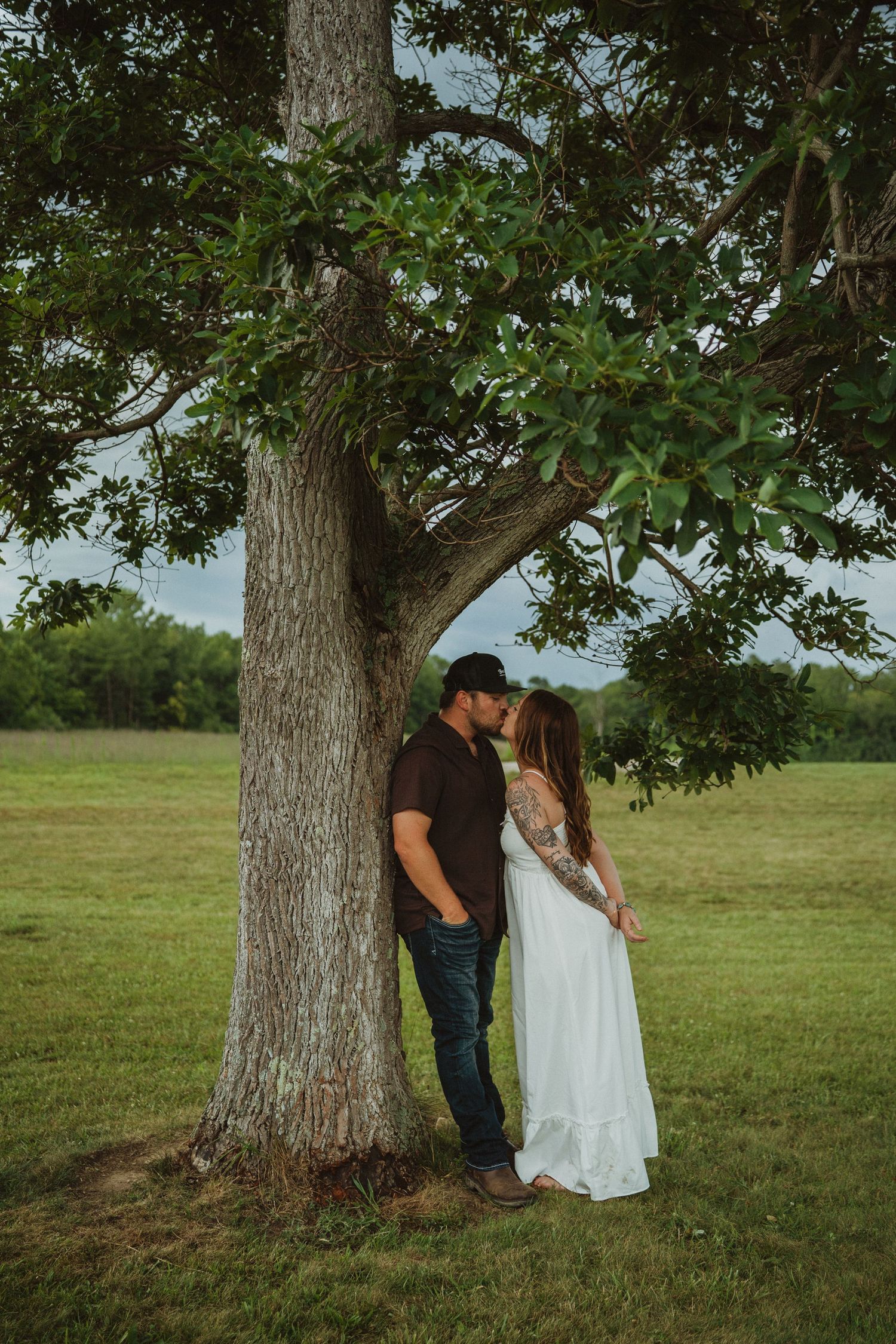 Couple sharing a romantic moment under a large oak tree, woman in white dress, man in dark shirt.