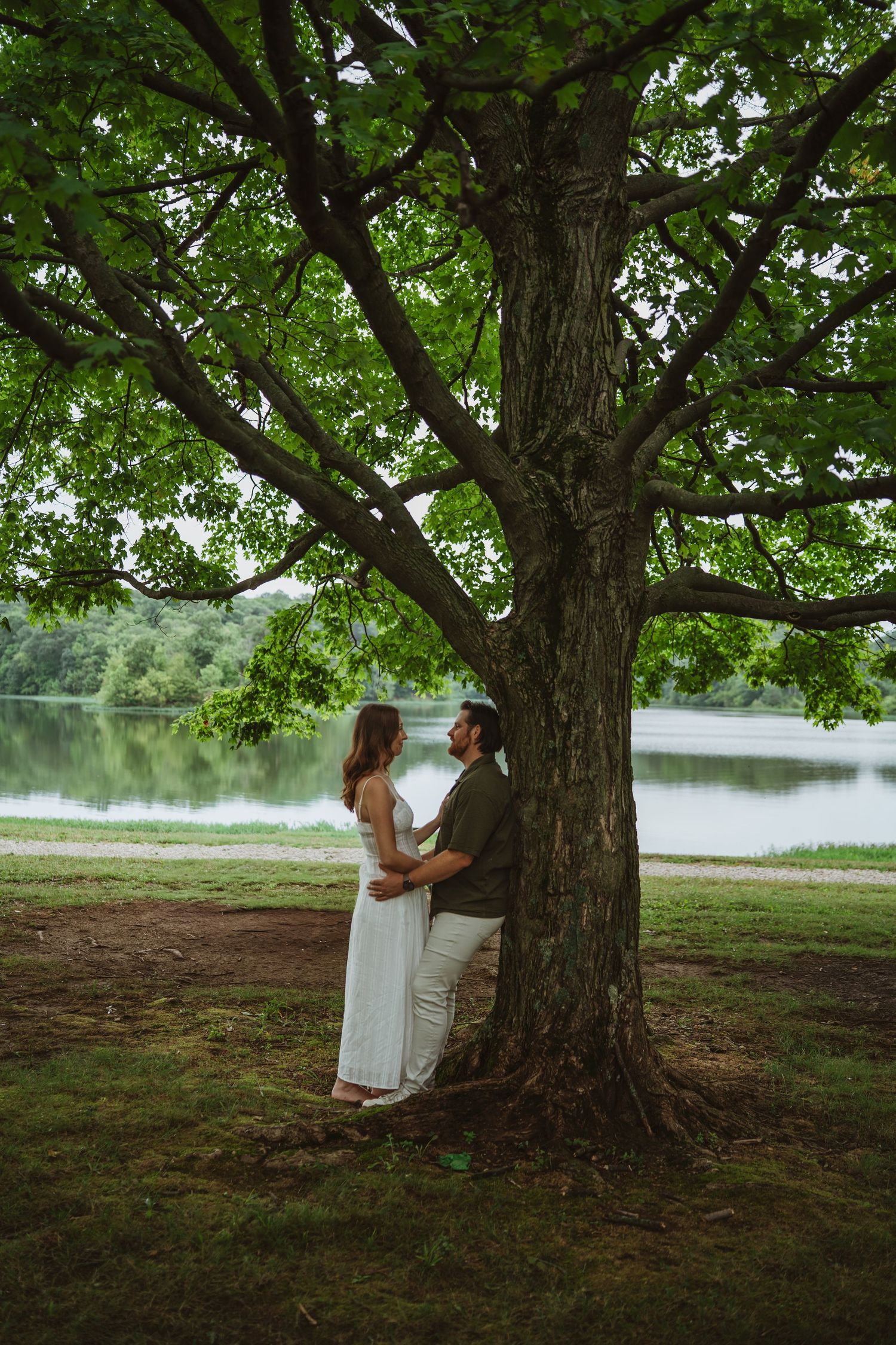 Couple sharing a tender moment under a large tree by a serene lake during a maternity photoshoot.