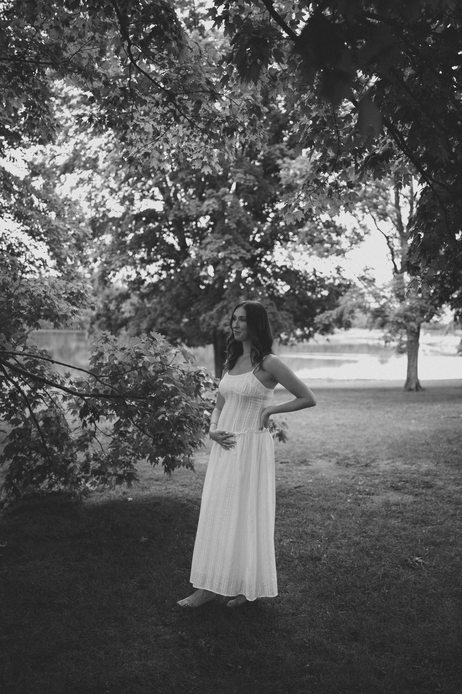 Pregnant woman in white maxi dress standing gracefully under trees in park, elegant black and white portrait.