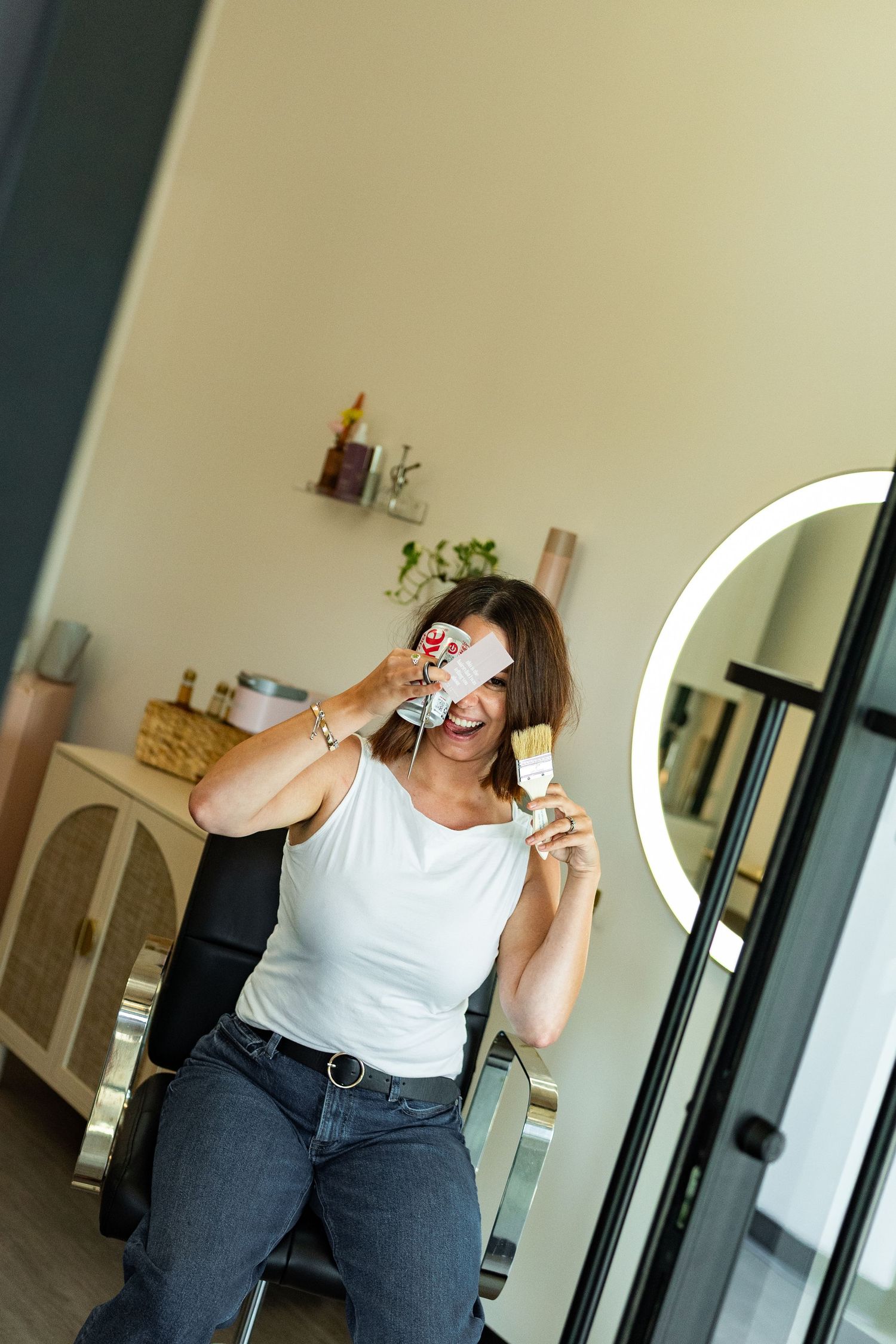 A cheerful woman in a white top holds a makeup brush and product, posing near a ring light in a beauty salon.