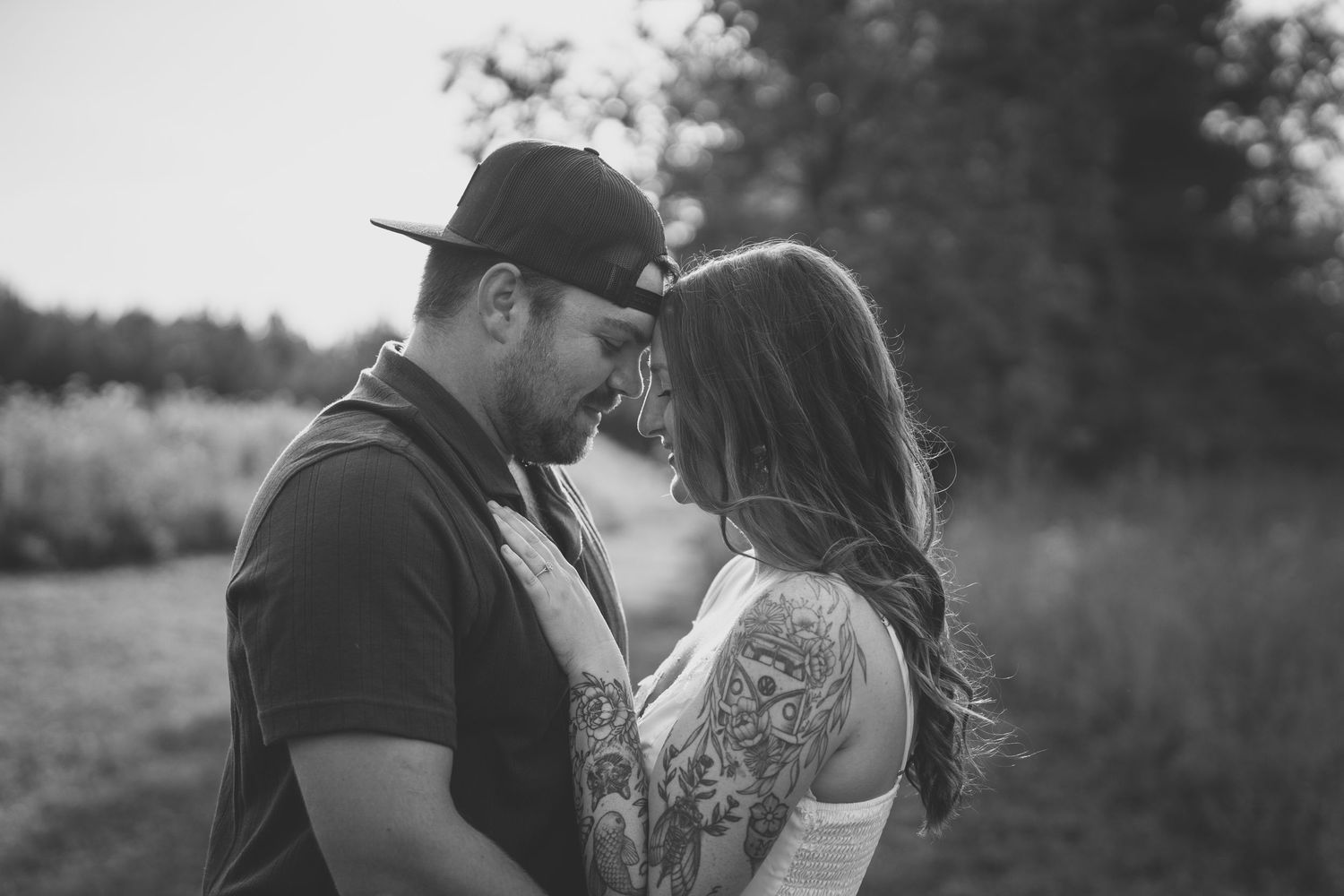 Black and white photo of a tattooed couple sharing an intimate moment outdoors, nearly kissing in a natural setting.