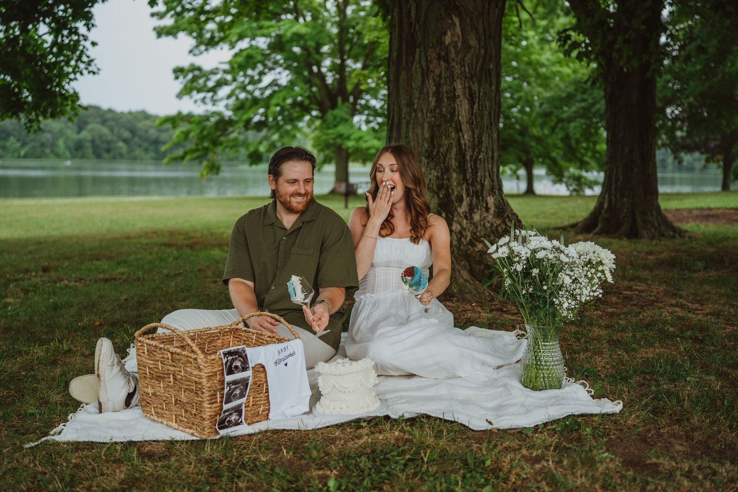 Couple sitting on white blanket under tree by lake, celebrating with champagne and picnic basket.