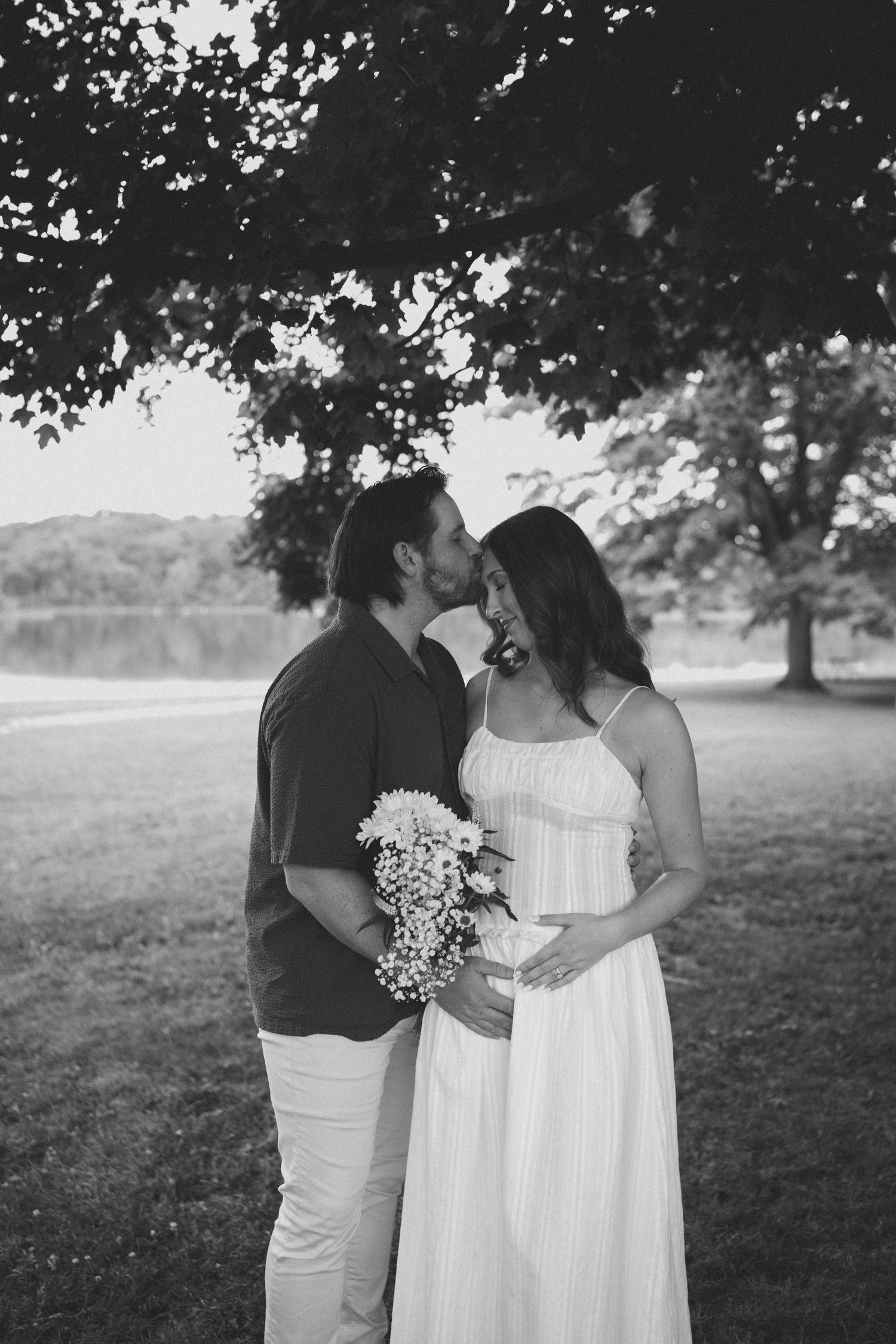 Black and white photo of a couple sharing a kiss outdoors near a lake, woman in white dress holding a floral bouquet.