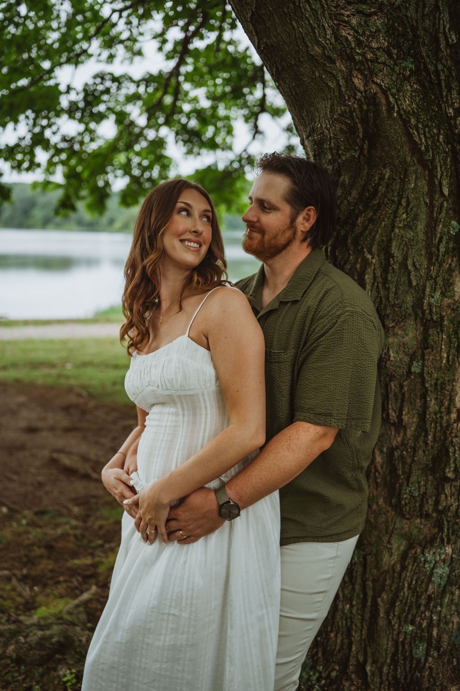 Couple embracing by a large tree near a lake, woman in white dress, man in green shirt, outdoor engagement photo.