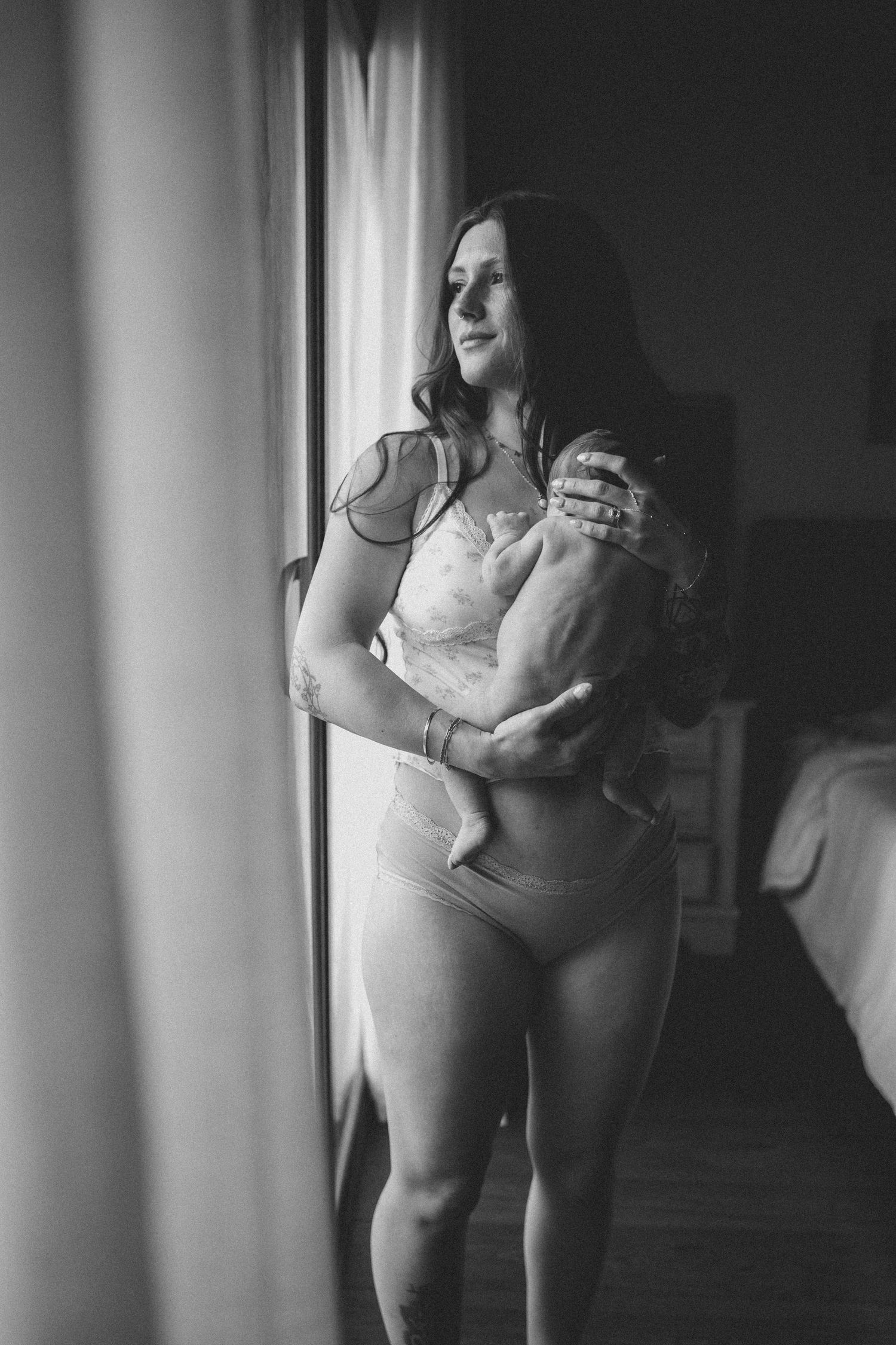 Black and white portrait of a woman in lingerie holding a newborn baby near a bright window.