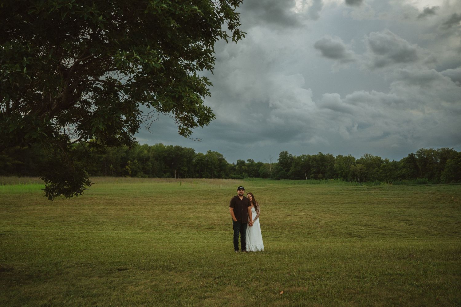 Couple embracing in a vast green field under dramatic stormy skies, surrounded by lush trees in a romantic outdoor setting.