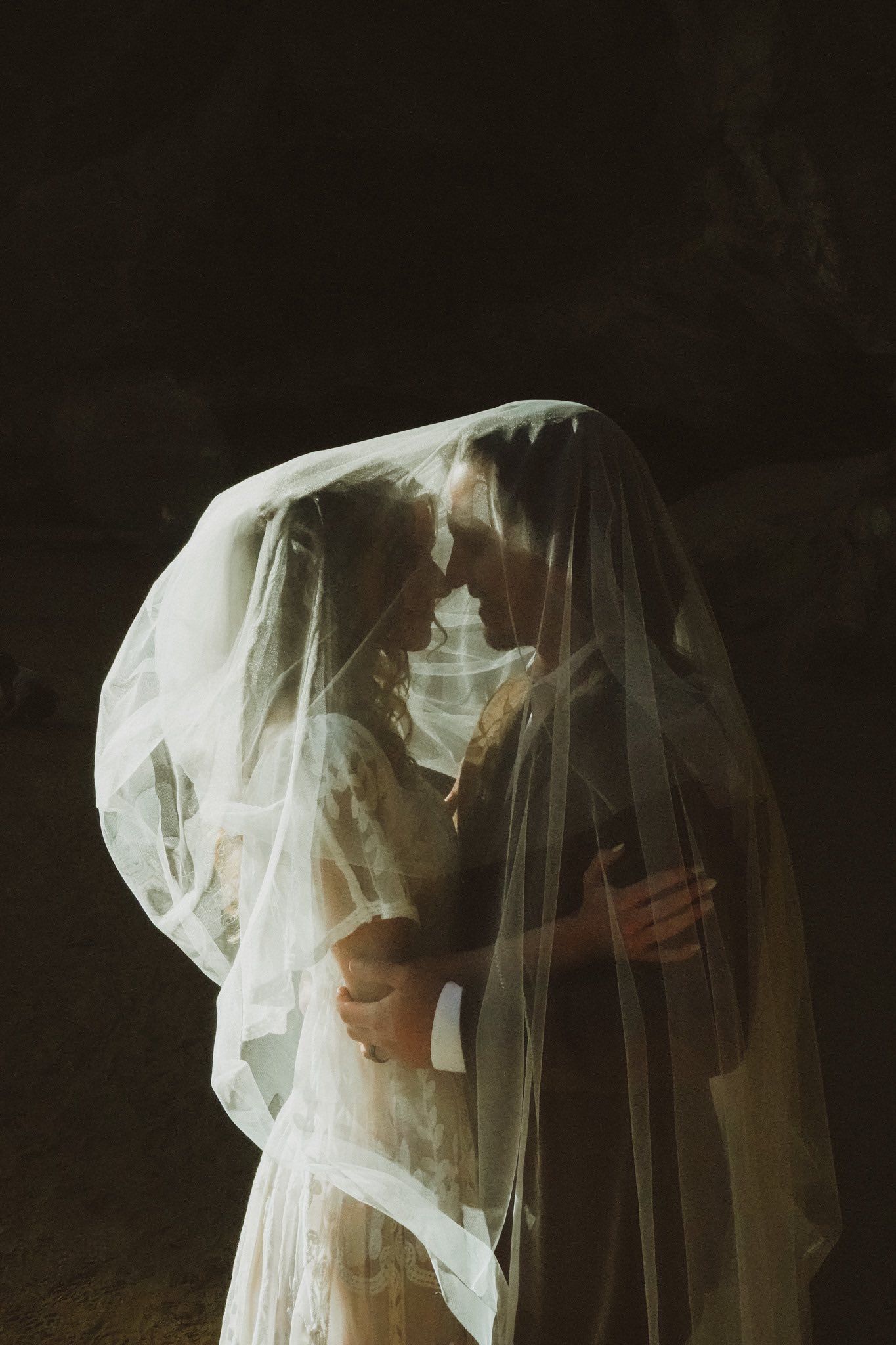 A romantic silhouette of a couple embracing under a flowing white wedding veil against a dark background.