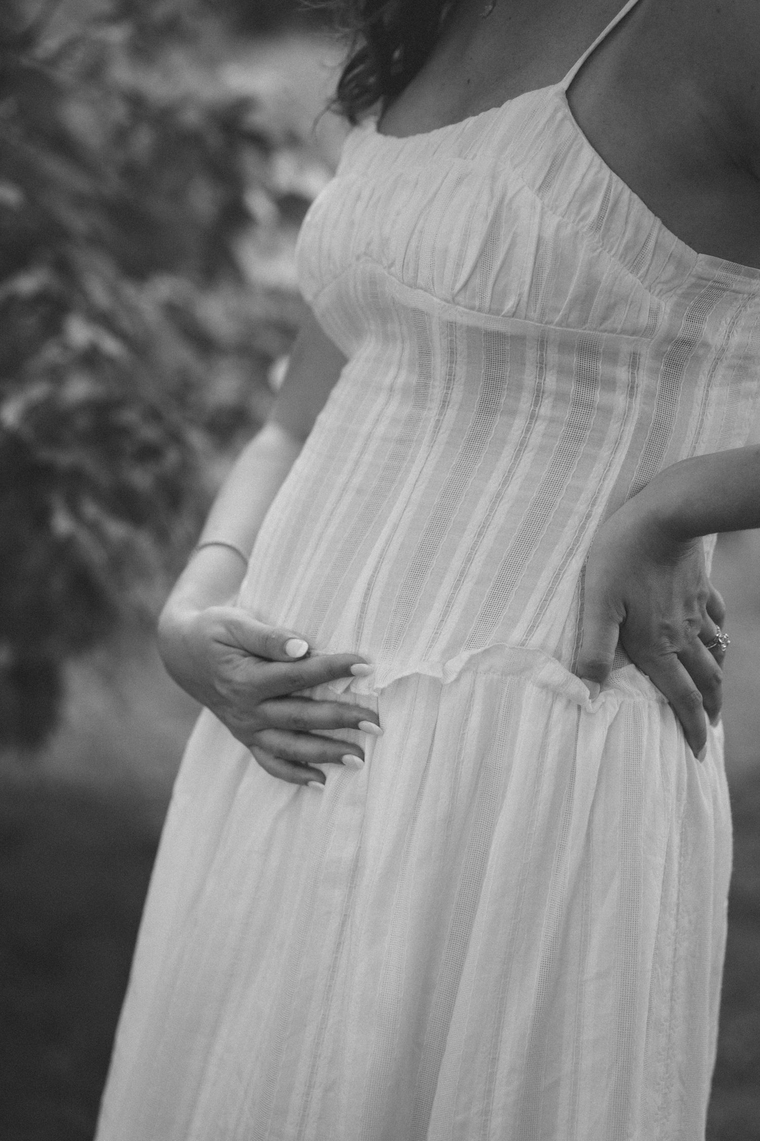 Black and white photo of a pregnant woman in a white dress holding her baby bump outdoors.