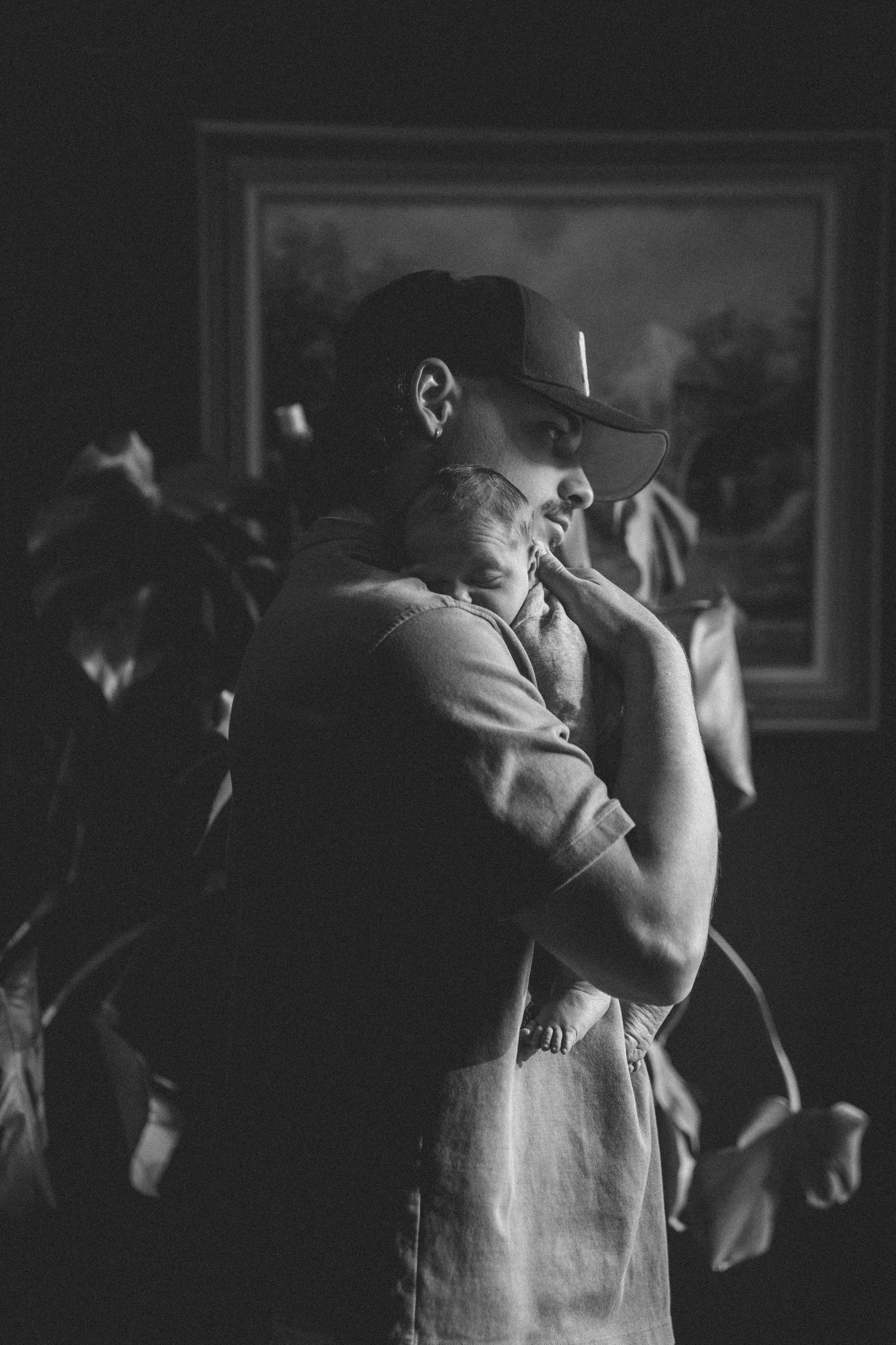 Black and white photo of a father tenderly holding his newborn baby close to his chest near a houseplant.