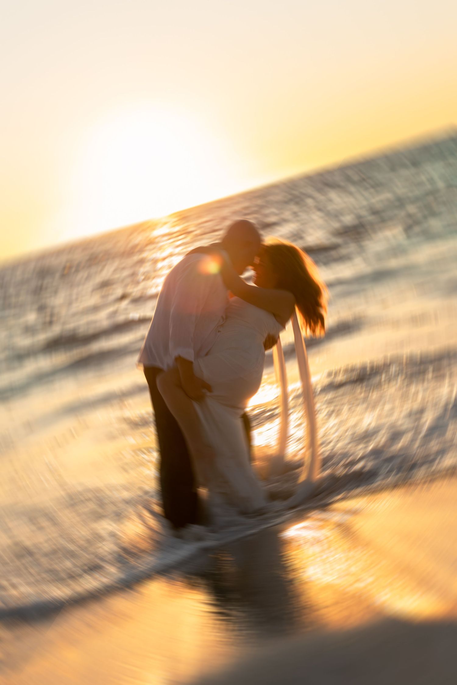 Romantic couple kissing on a beach at golden sunset, waves splashing around them in a dreamy, blurred motion.