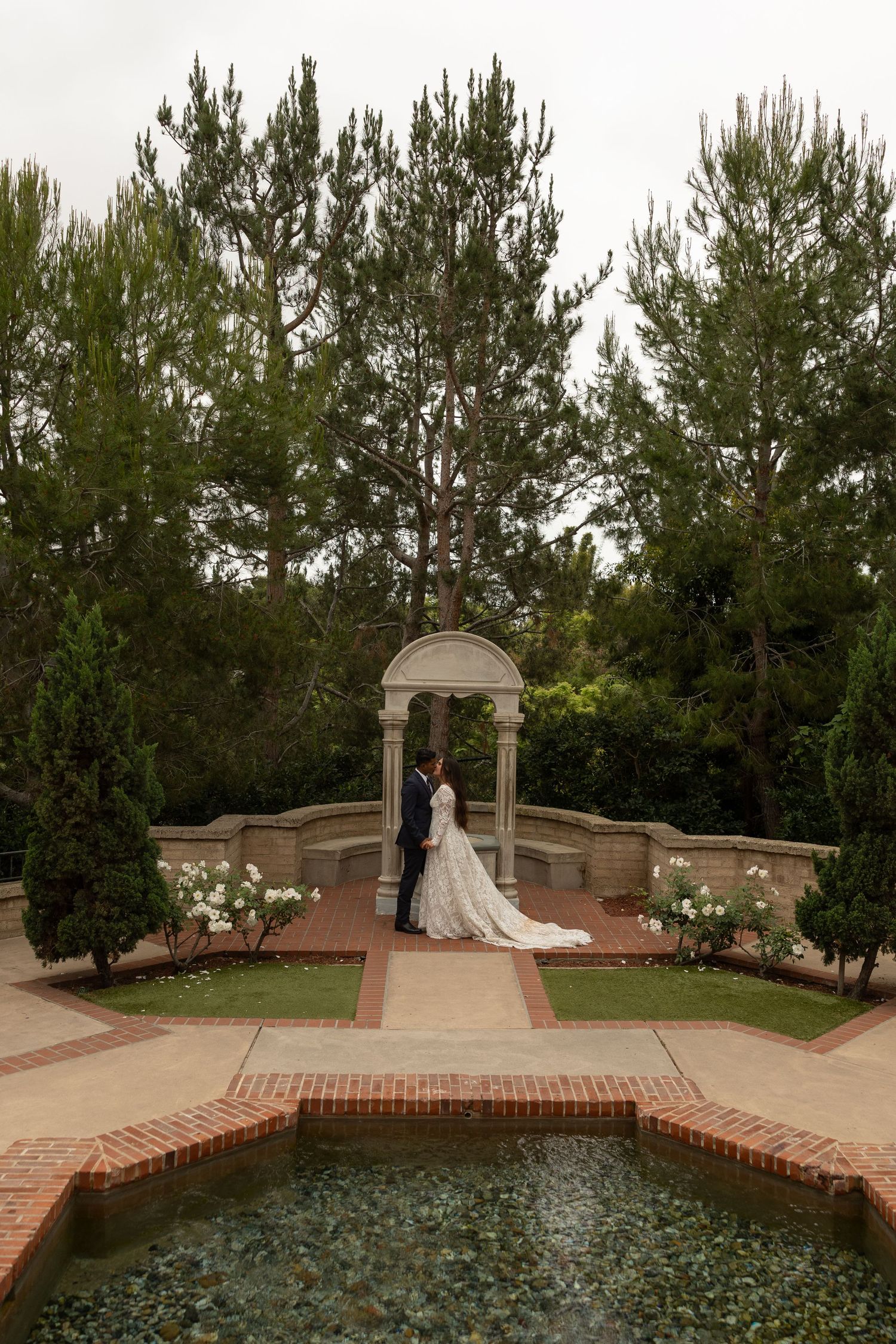 Bride and groom stand beneath stone arch gazebo surrounded by lush gardens and reflecting pool at elegant outdoor wedding.
