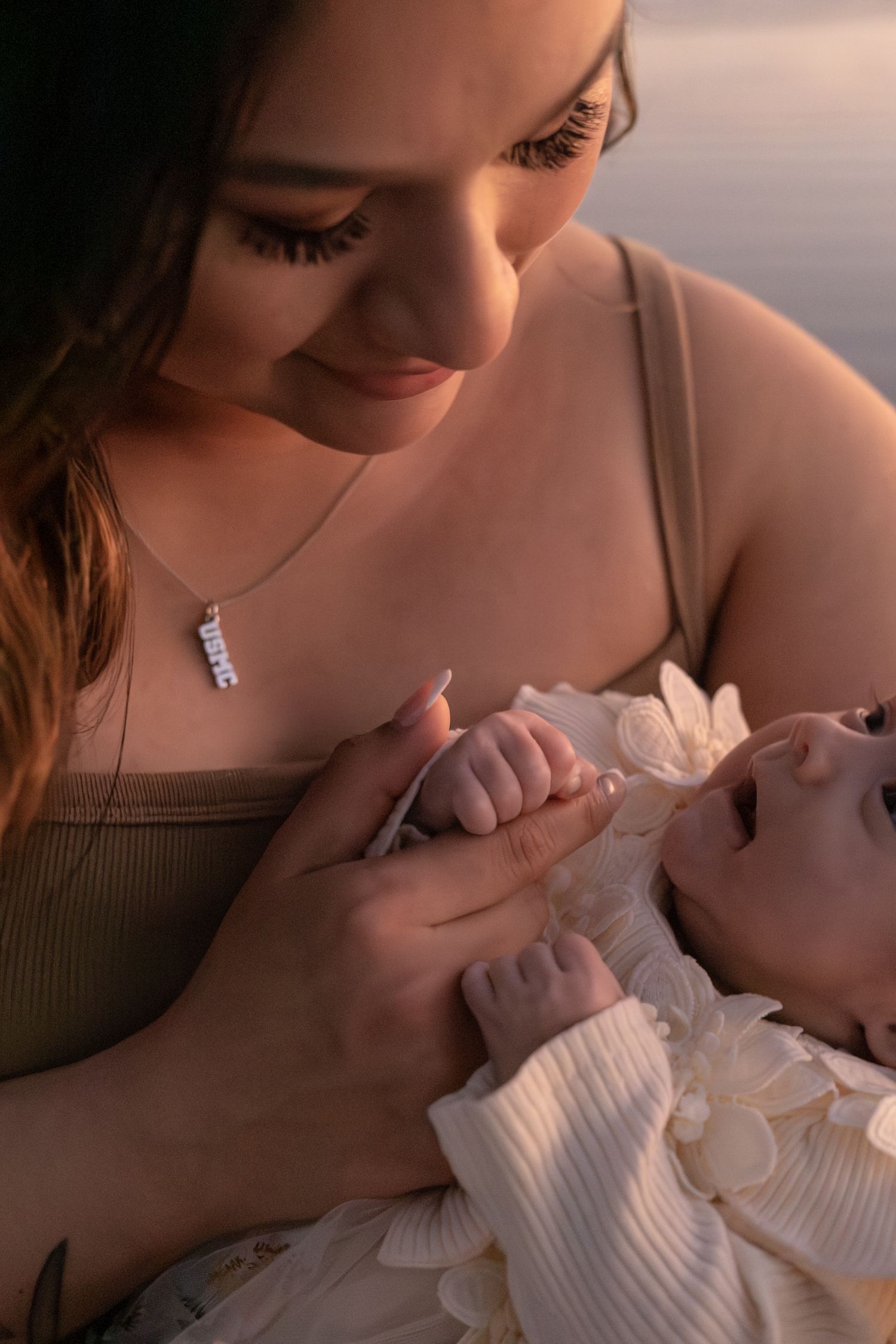 Young mother with necklace smiling down at her newborn baby in warm golden light.