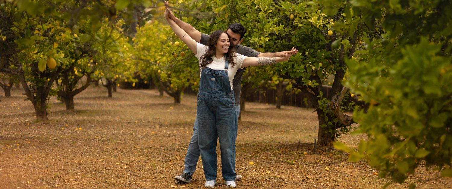 A joyful couple embraces in a citrus orchard, woman in denim overalls with arms outstretched.