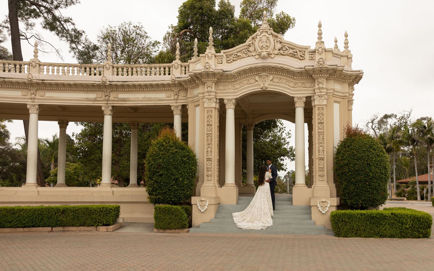 Bride in white wedding gown standing on steps of ornate classical pavilion with columns at Balboa Park, San Diego.