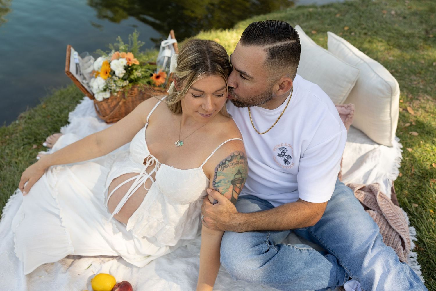 A couple shares a romantic outdoor picnic by a lake, with the man kissing the woman's cheek as she leans against him.
