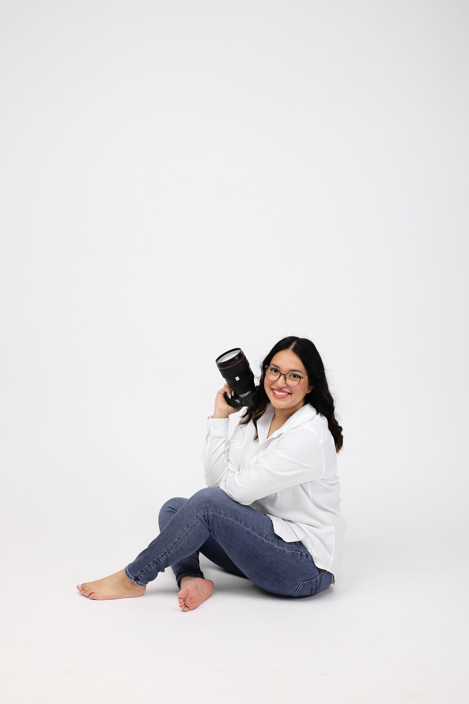 A smiling woman with glasses sits on a white floor holding a camera lens, wearing a white shirt and jeans.