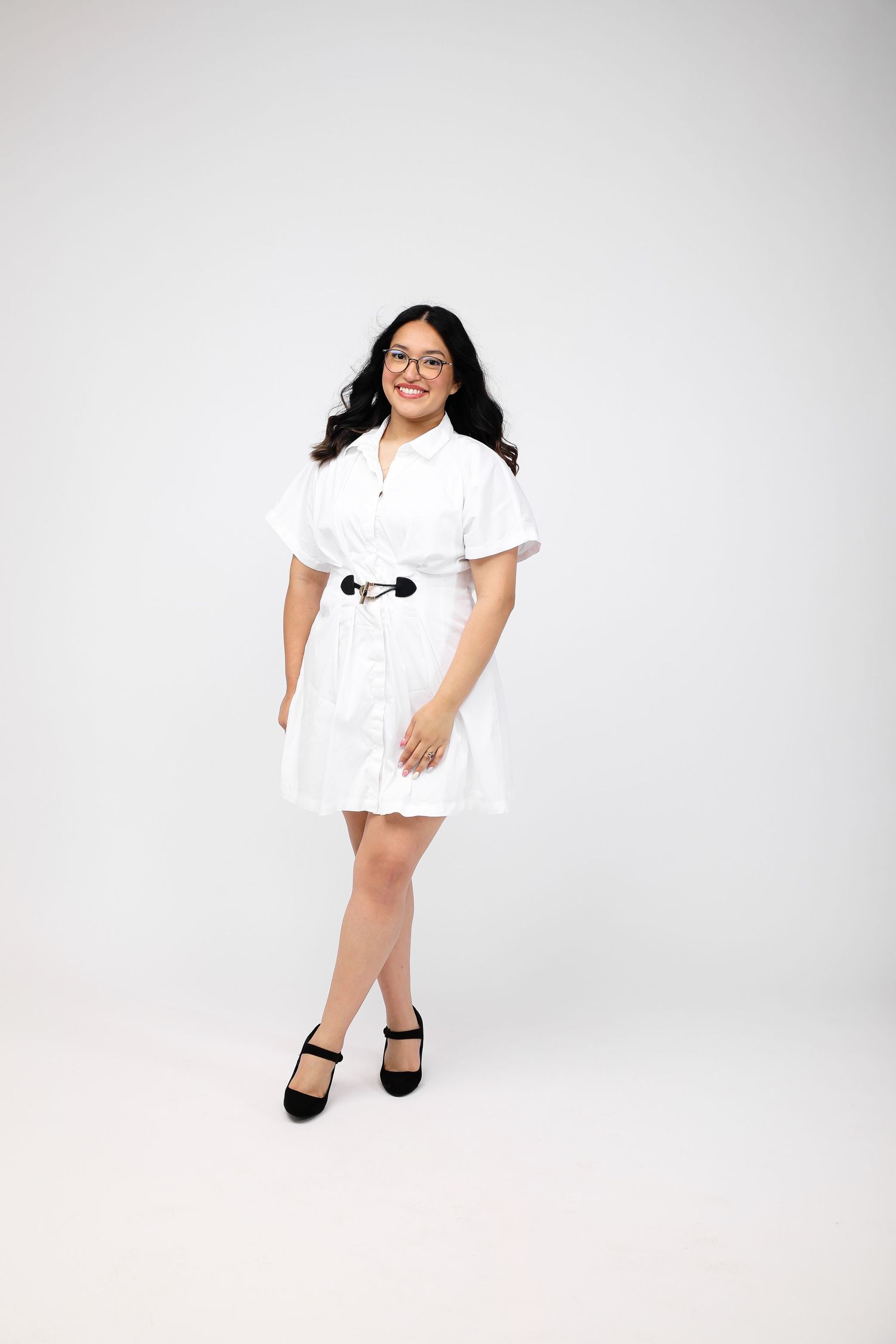 Young woman in white belted mini dress with short sleeves, black heels, and glasses, posing on white background.