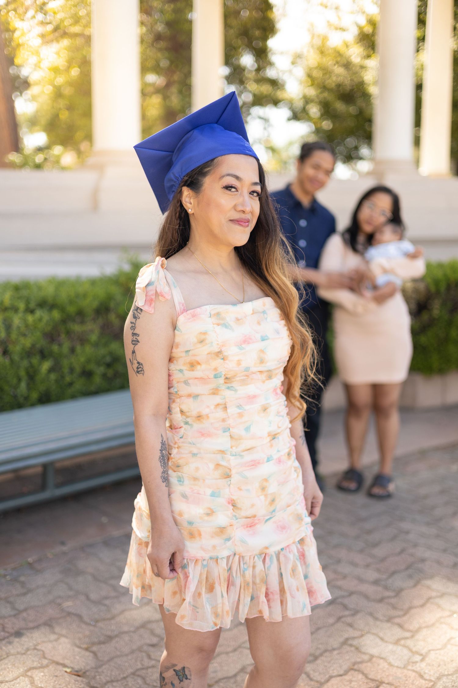 A woman in a floral dress and blue graduation cap smiles while family members stand in the background.