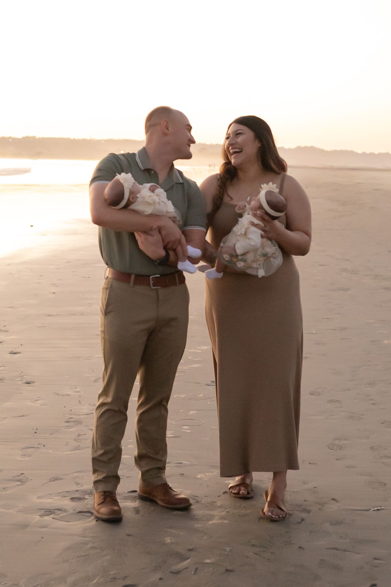 Couple laughing together on a beach at sunset while each holding a newborn baby.