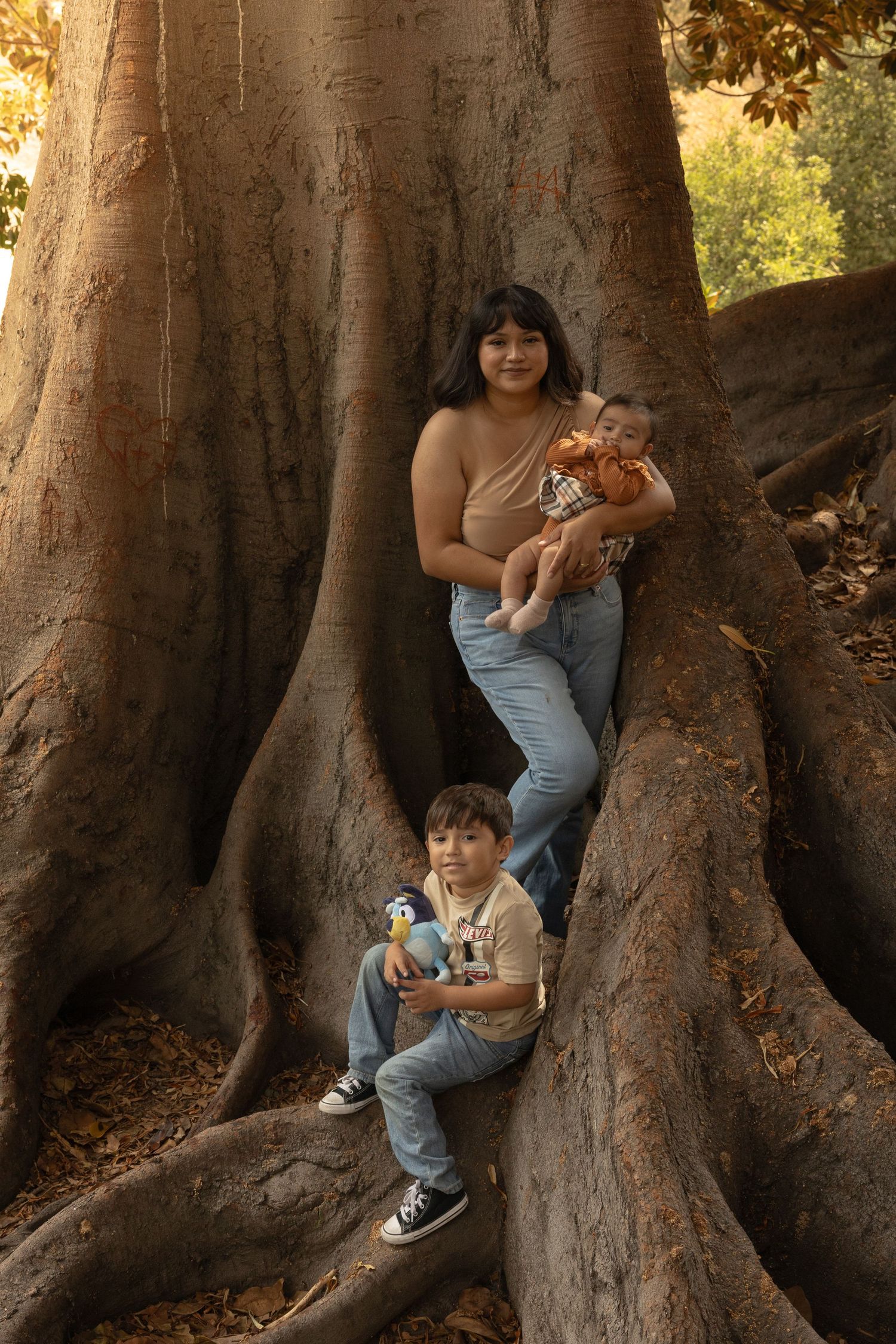 Young mother holding a baby with a toddler sitting among the roots of a large tree.