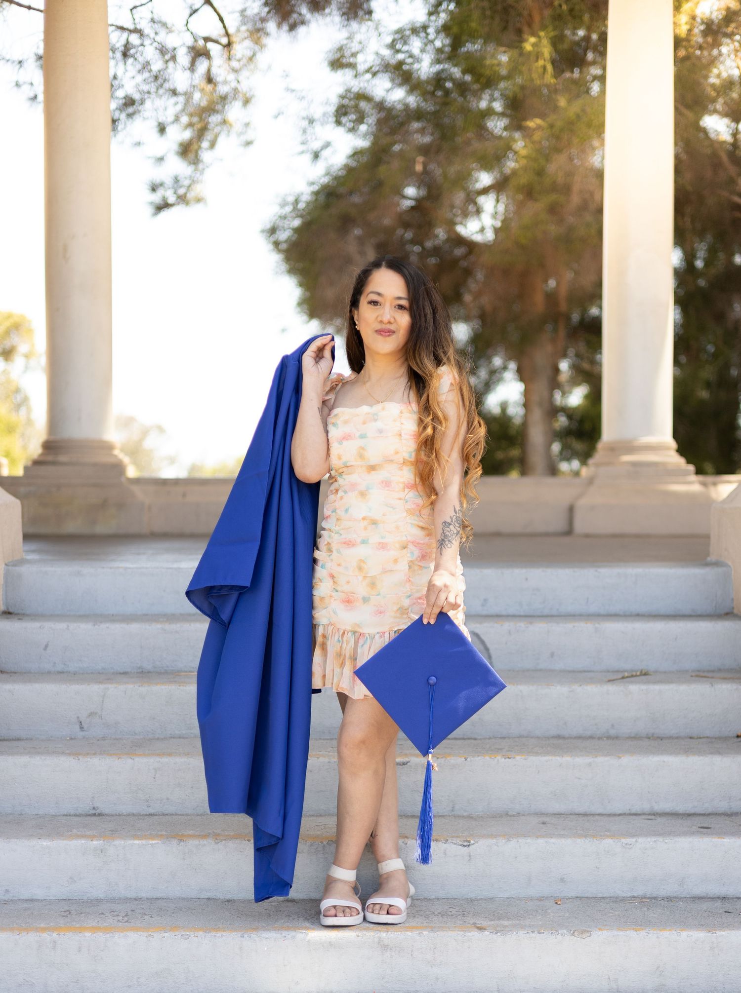 A graduate in a blue cap and gown poses on stone steps holding her cap and gown in a floral dress.