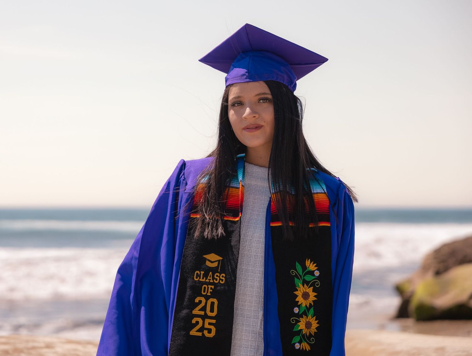 A graduate in a blue cap and gown wearing a Class of 2025 stole poses on a rocky beach.
