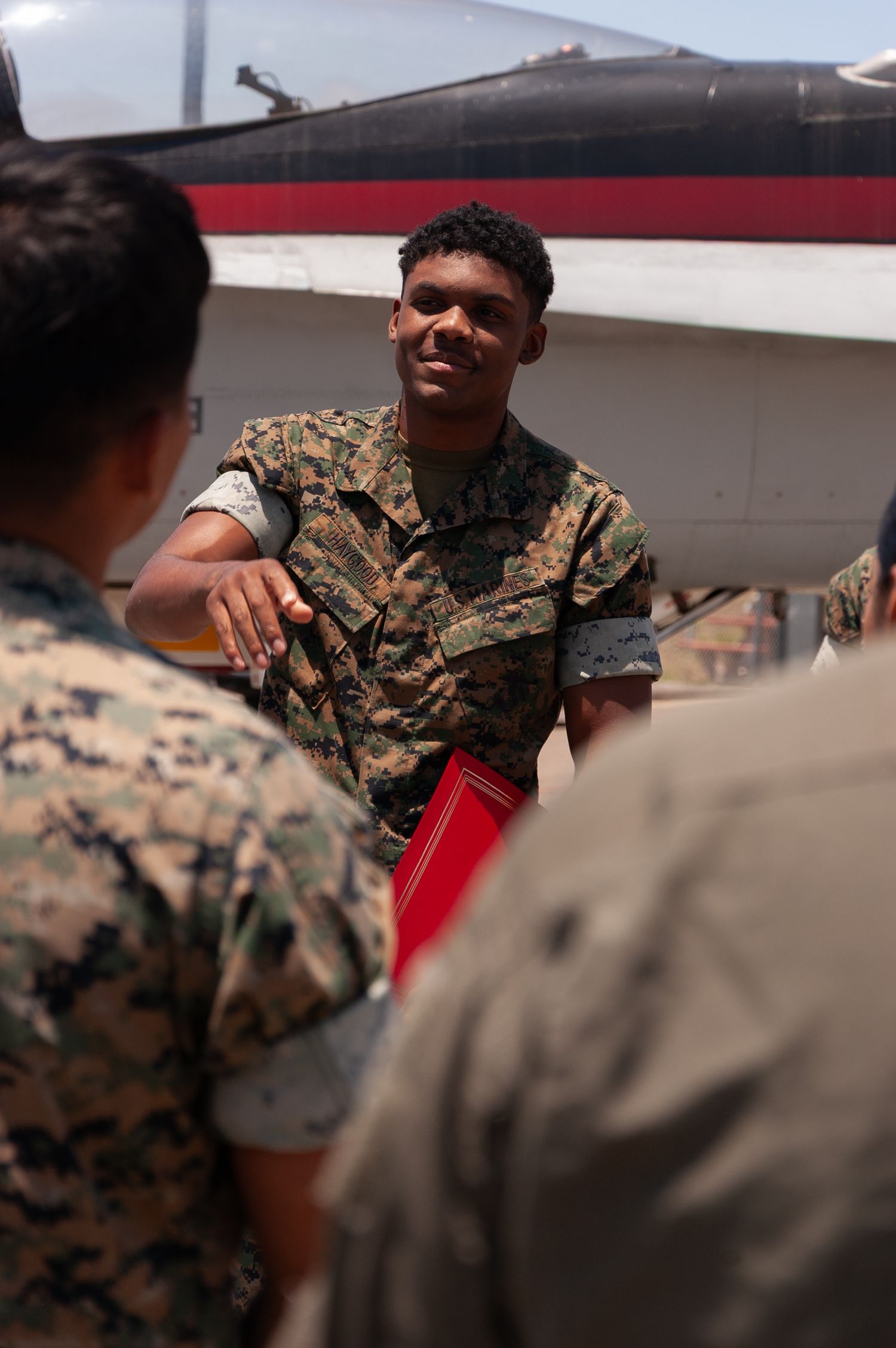 US Marine in camouflage uniform speaks to group near military aircraft on sunny tarmac.