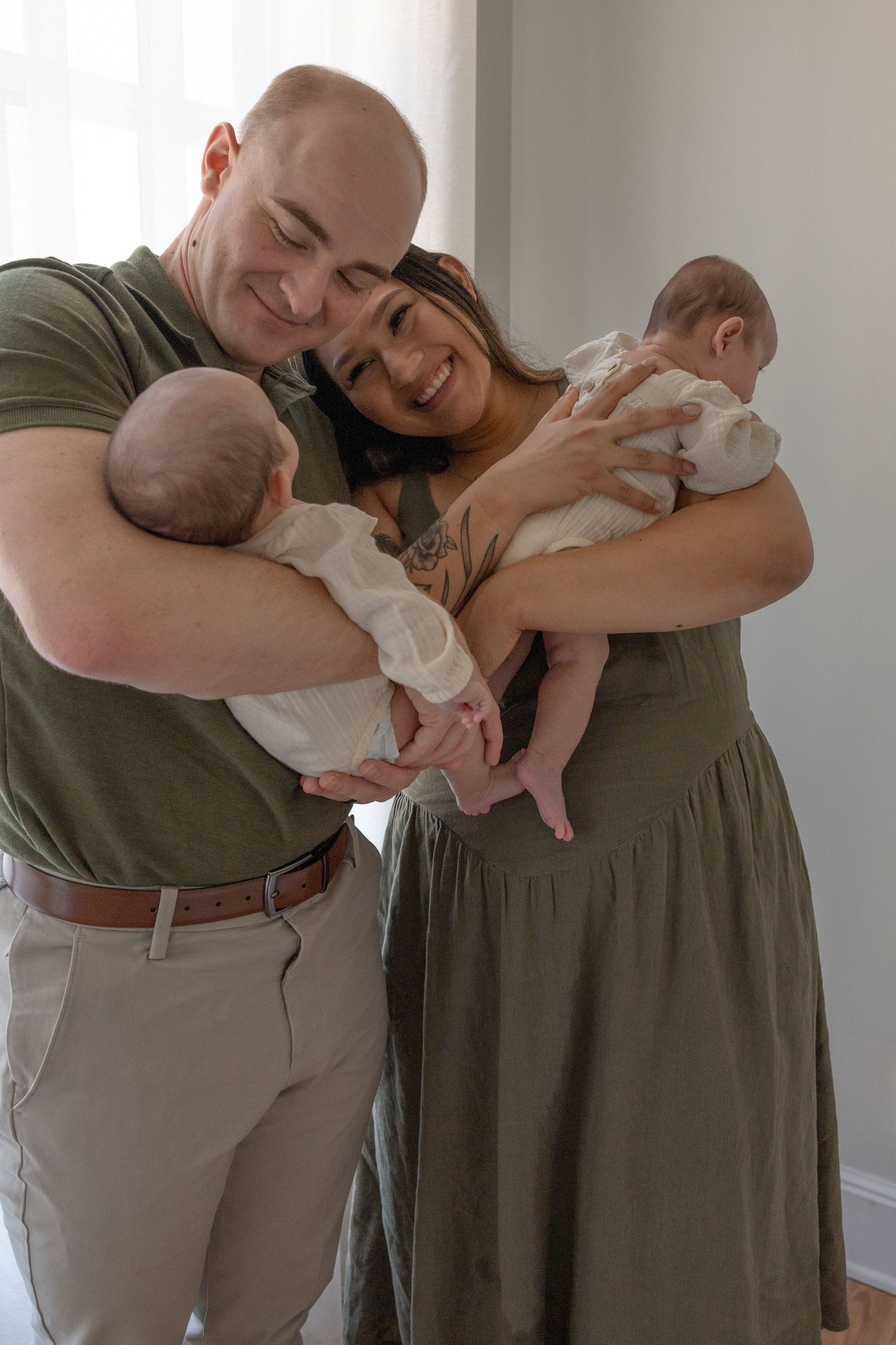 A happy couple holds their twin newborn babies while standing in a bright indoor setting.