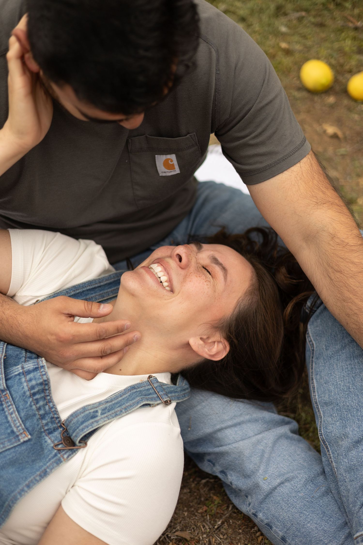 A laughing woman in denim overalls lies on a mans lap outdoors near citrus trees.