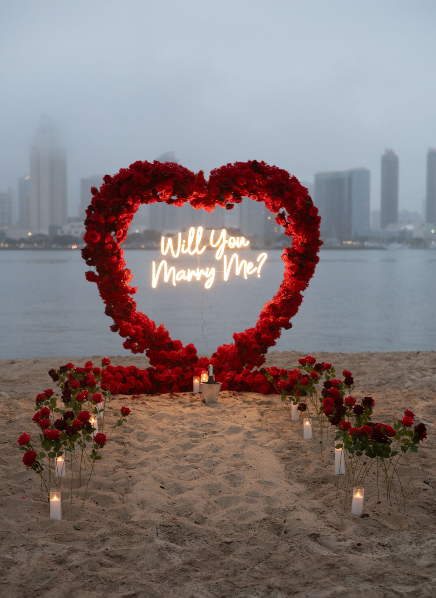 Romantic beach proposal setup with red rose heart arch and Will You Marry Me neon sign.