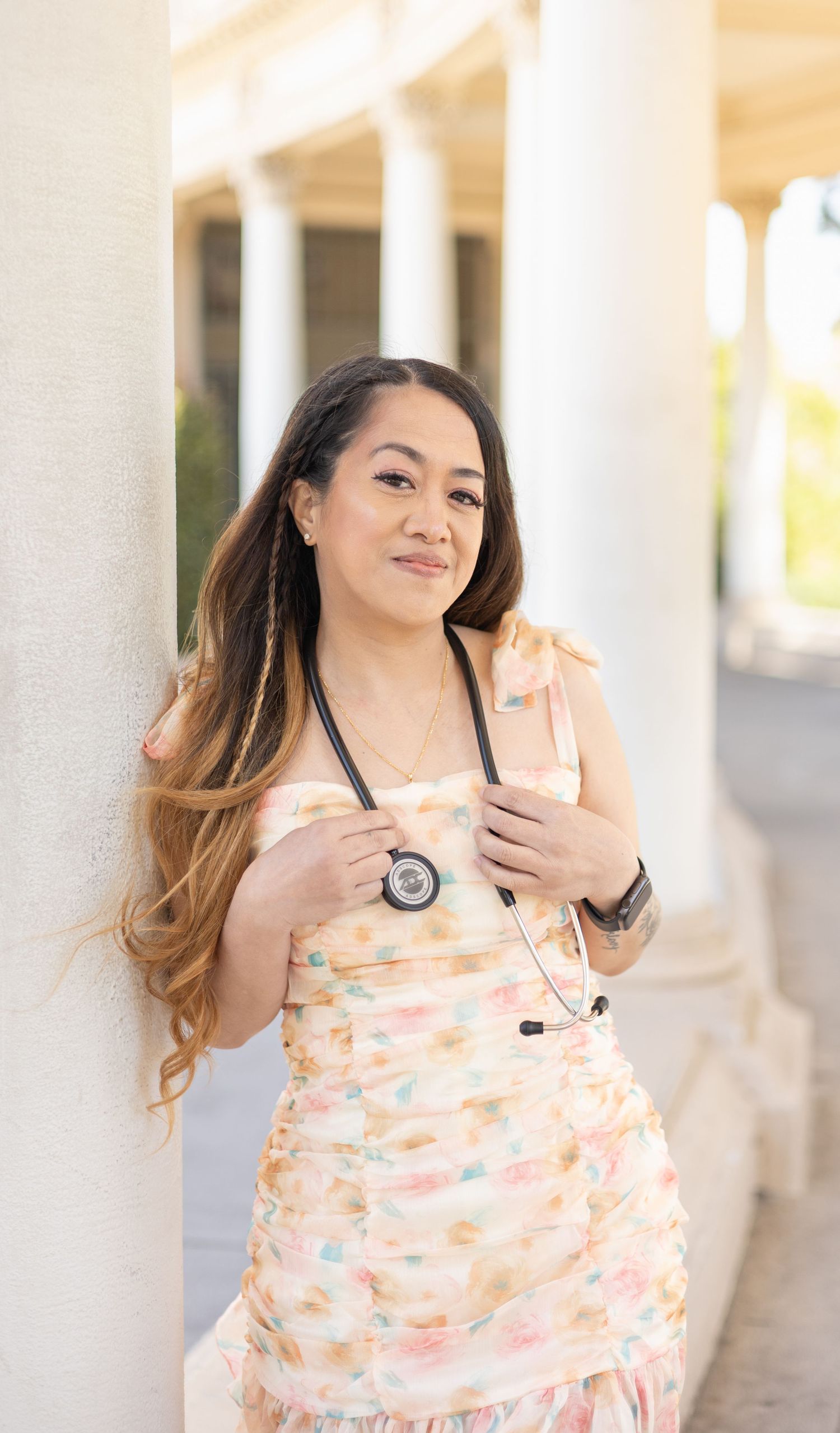 A woman in a floral dress wearing a stethoscope leans against a white column, smiling warmly.
