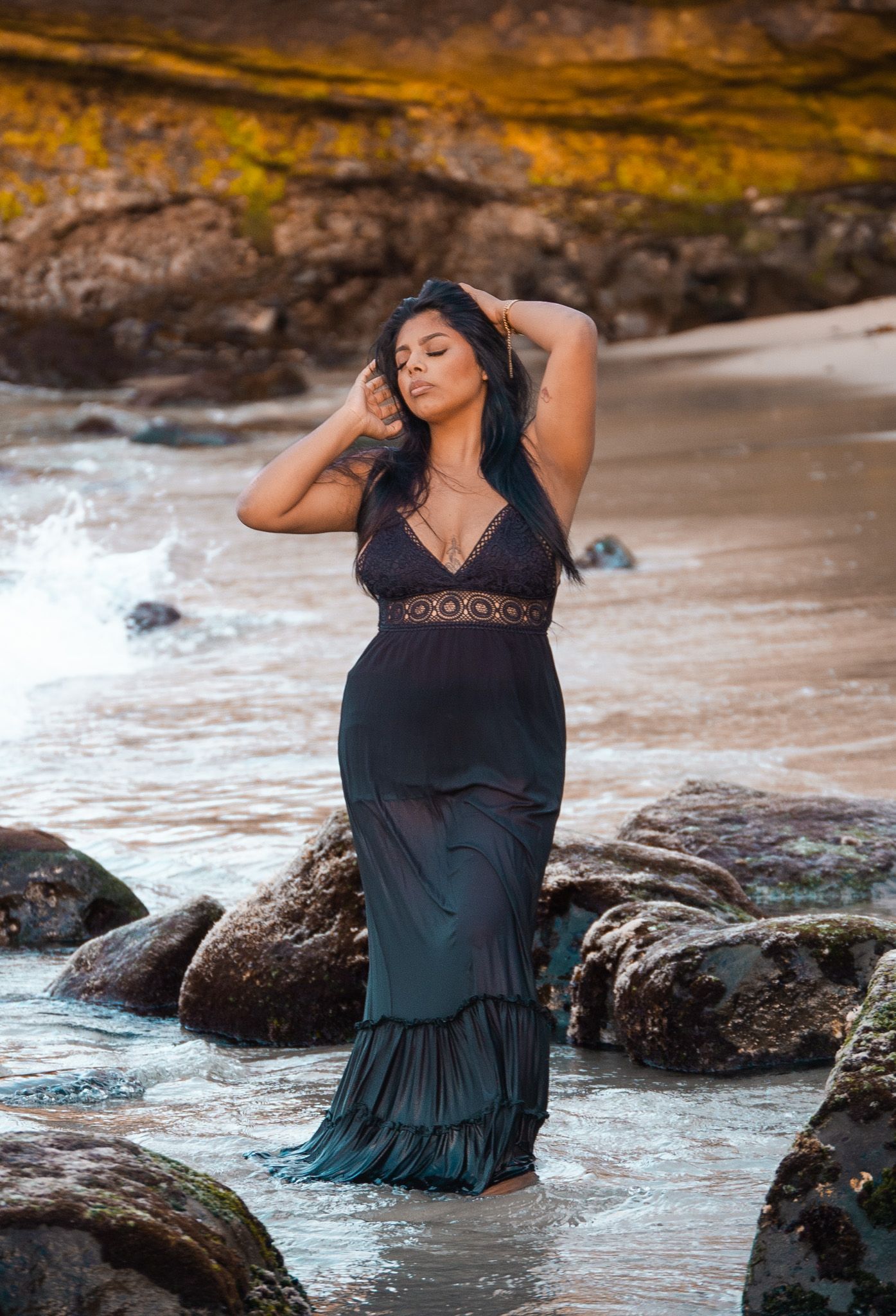 Woman in black lace maxi dress standing in ocean water among rocks at sunset.