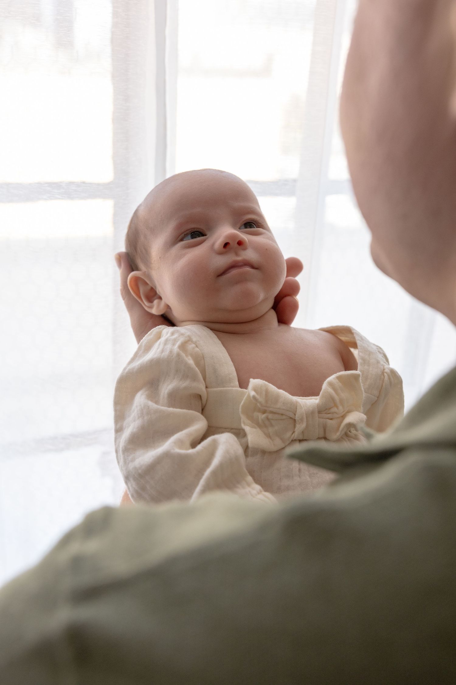 A newborn baby in a cream bow outfit being held up by an adult near a bright window.