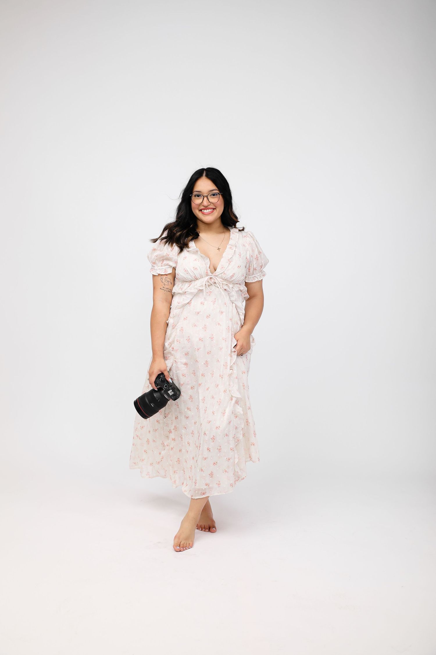 Woman in floral midi dress holding camera, smiling, barefoot on white background.
