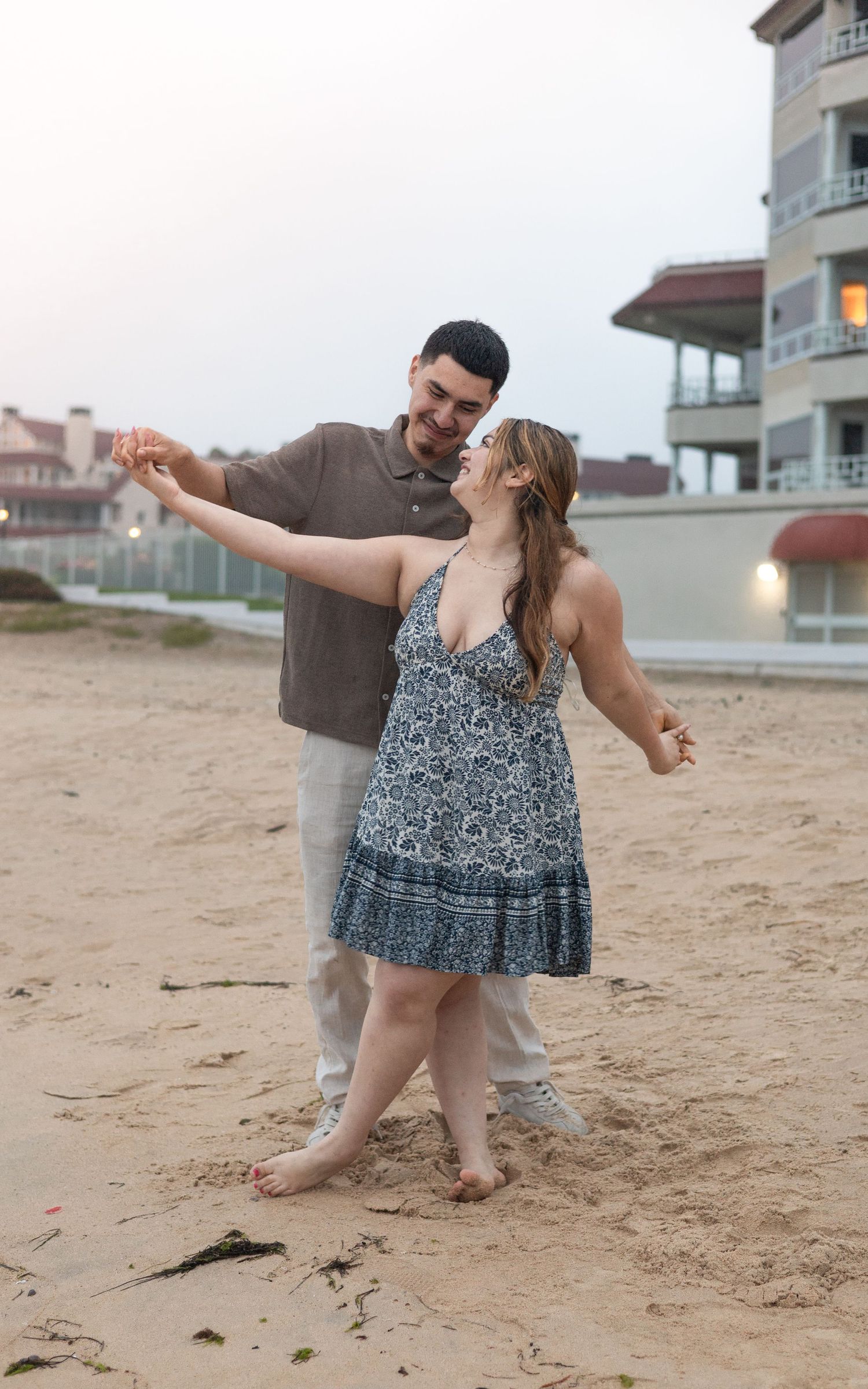 A happy couple dances barefoot on a sandy beach at dusk near a coastal building.